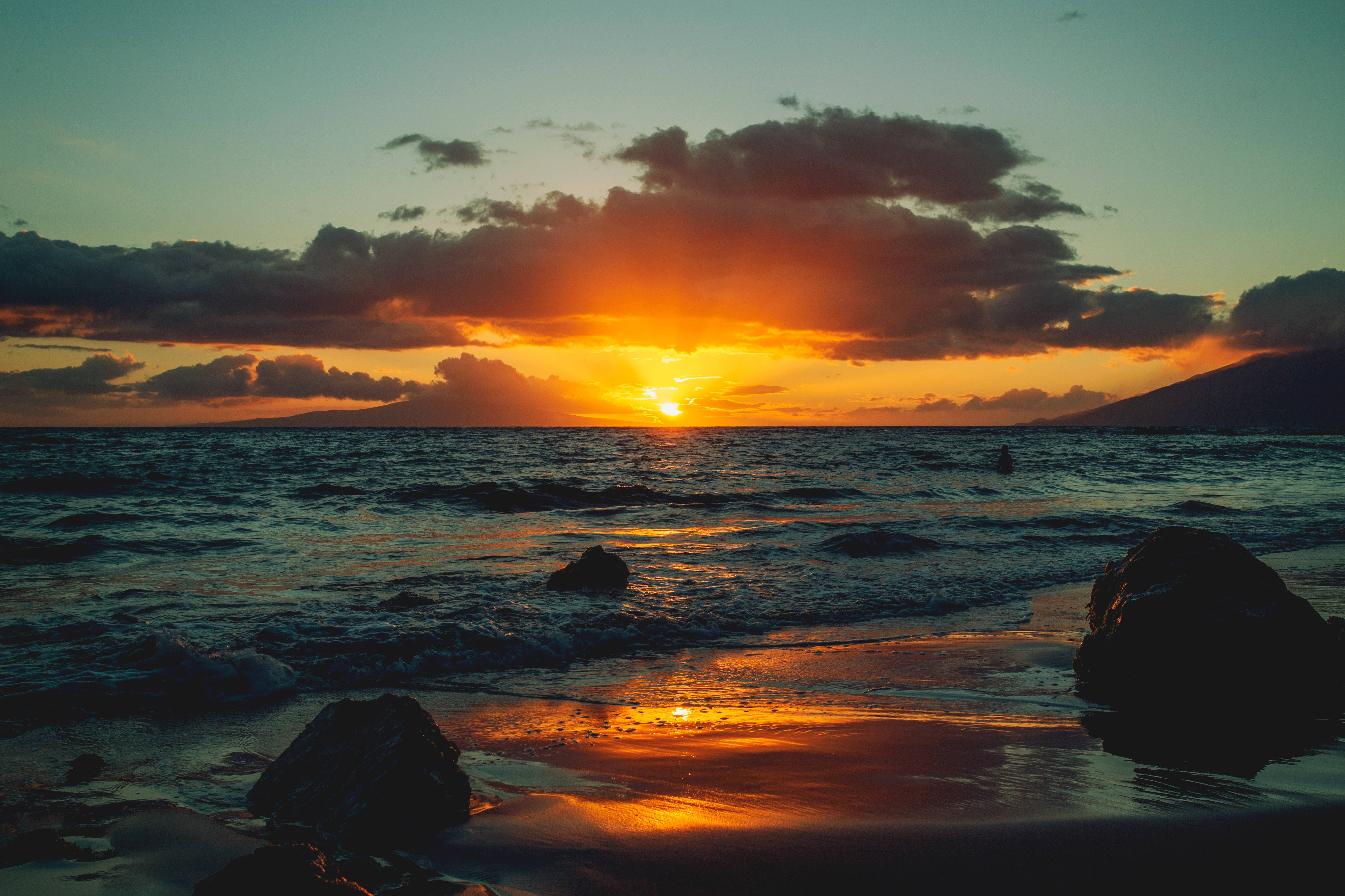 海洋 桌布 壁紙 — 熾熱的夕陽將海洋染成橙色和金色，海浪拍打著寧靜海灘上的黑色岩石。