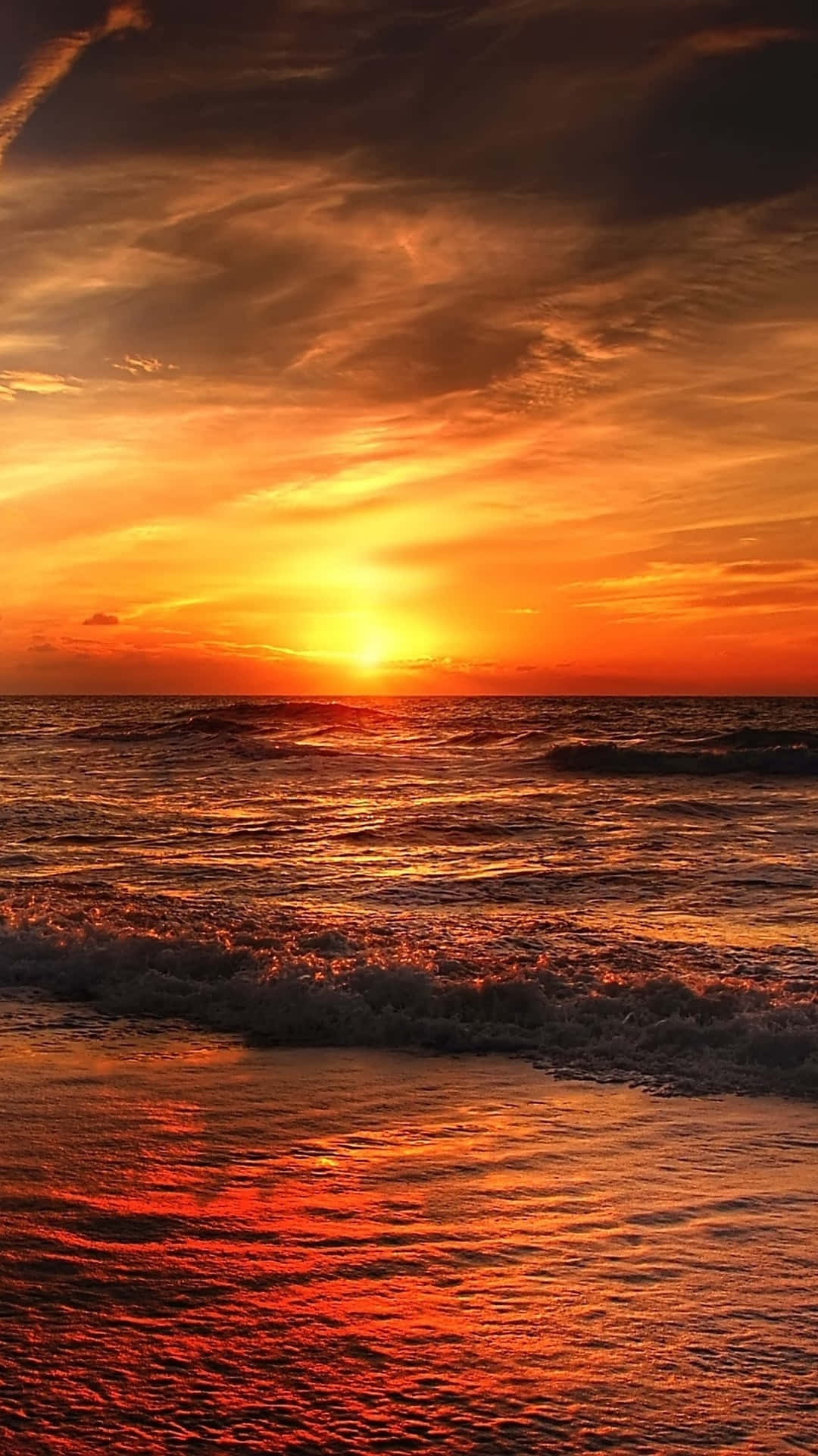 Hi Res Beach 桌布 壁紙 — 熾熱的夕陽將海洋染成發光的橙色和紅色，海浪在戲劇性的天空下拍打海岸。