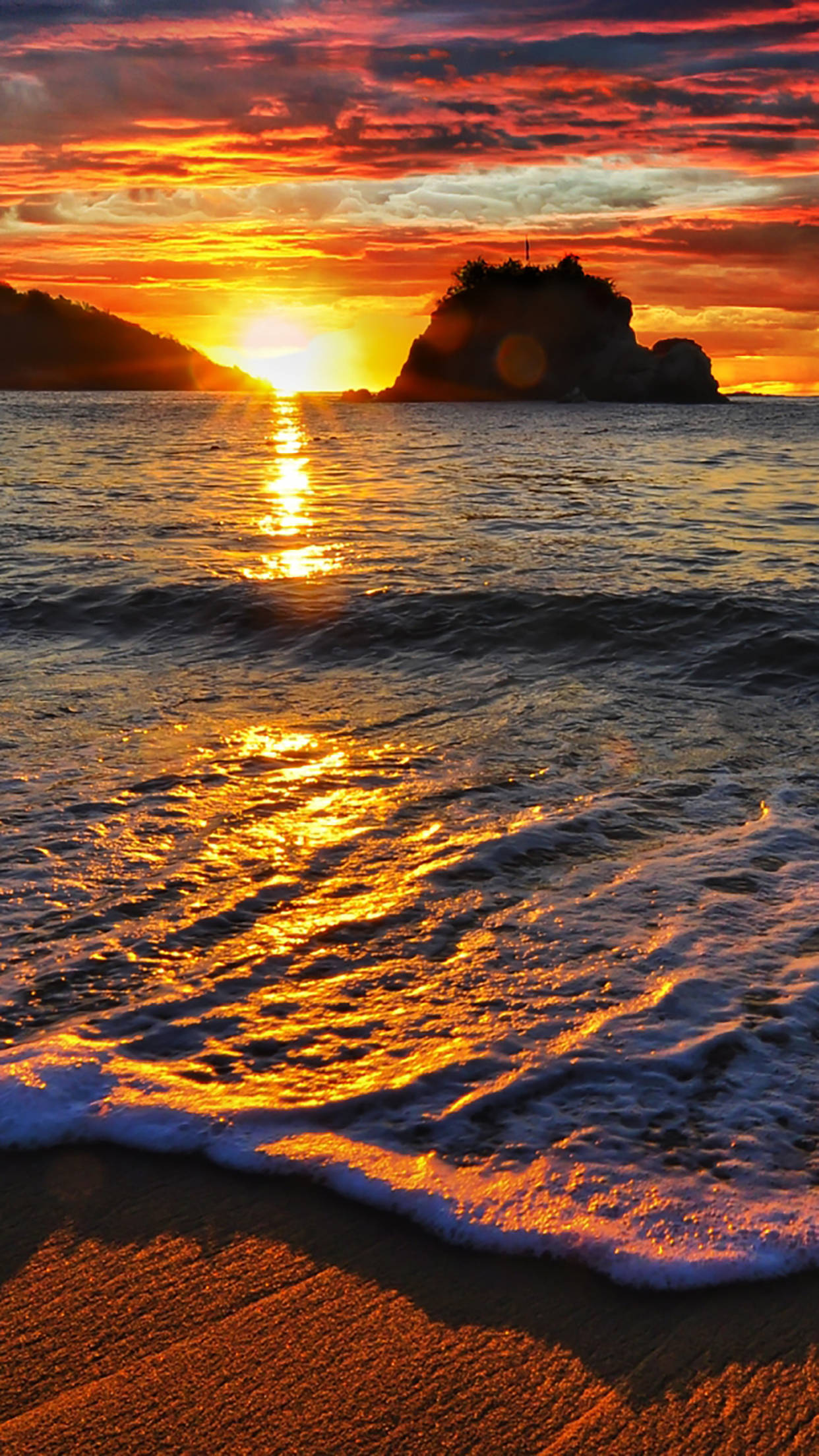 海灘日落 桌布 壁紙 — 熾熱的夕陽將海洋染成金色，海浪拍打著沙灘，旁邊是岩石島的剪影。