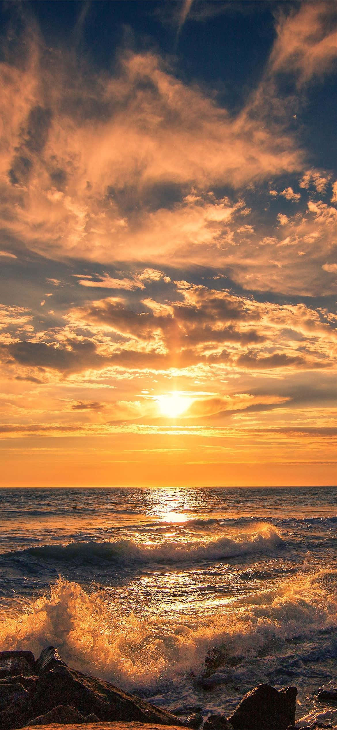 Surfing iPhone 桌布 壁紙 — 熾熱的夕陽用金色光芒描繪海洋，海浪在戲劇性的雲層下拍打著黑暗的岩石。