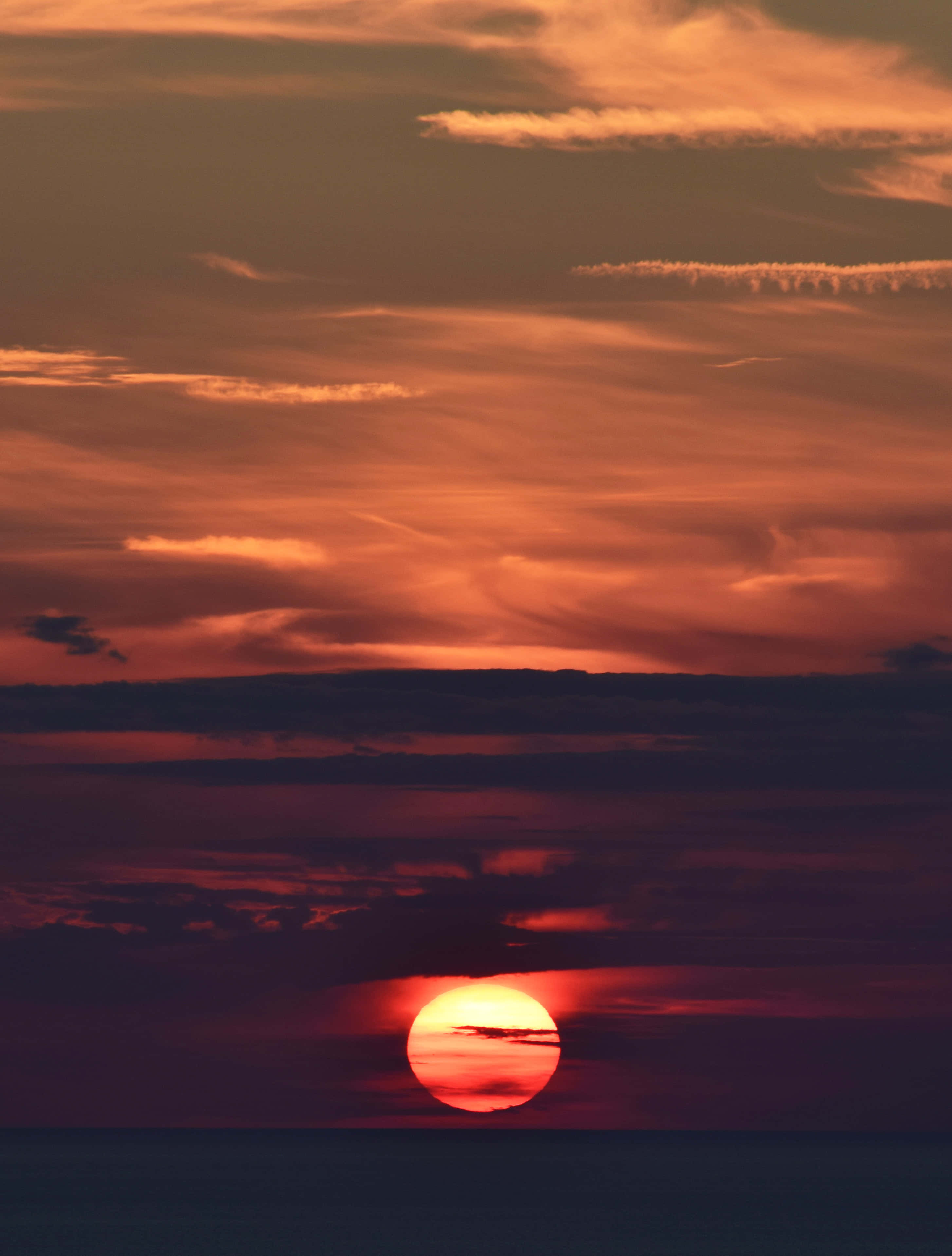 Sunset iPhone 桌布 壁紙 — 熾熱的太陽沉入地平線，在平靜的海洋上空，將鮮豔的橙色和深紫色色調投射在紋理豐富的天空中。