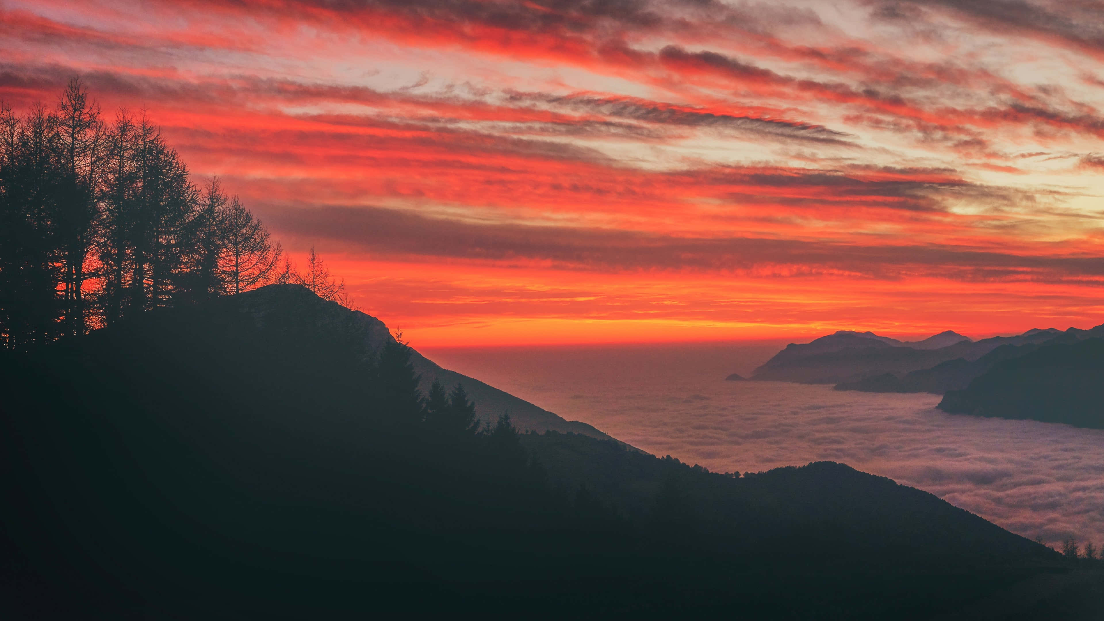 Mountain Aesthetic 桌布 壁紙 — 熾熱的橙紅色日落描繪了天空，下方是夾在黑暗山脈輪廓之間的雲海。