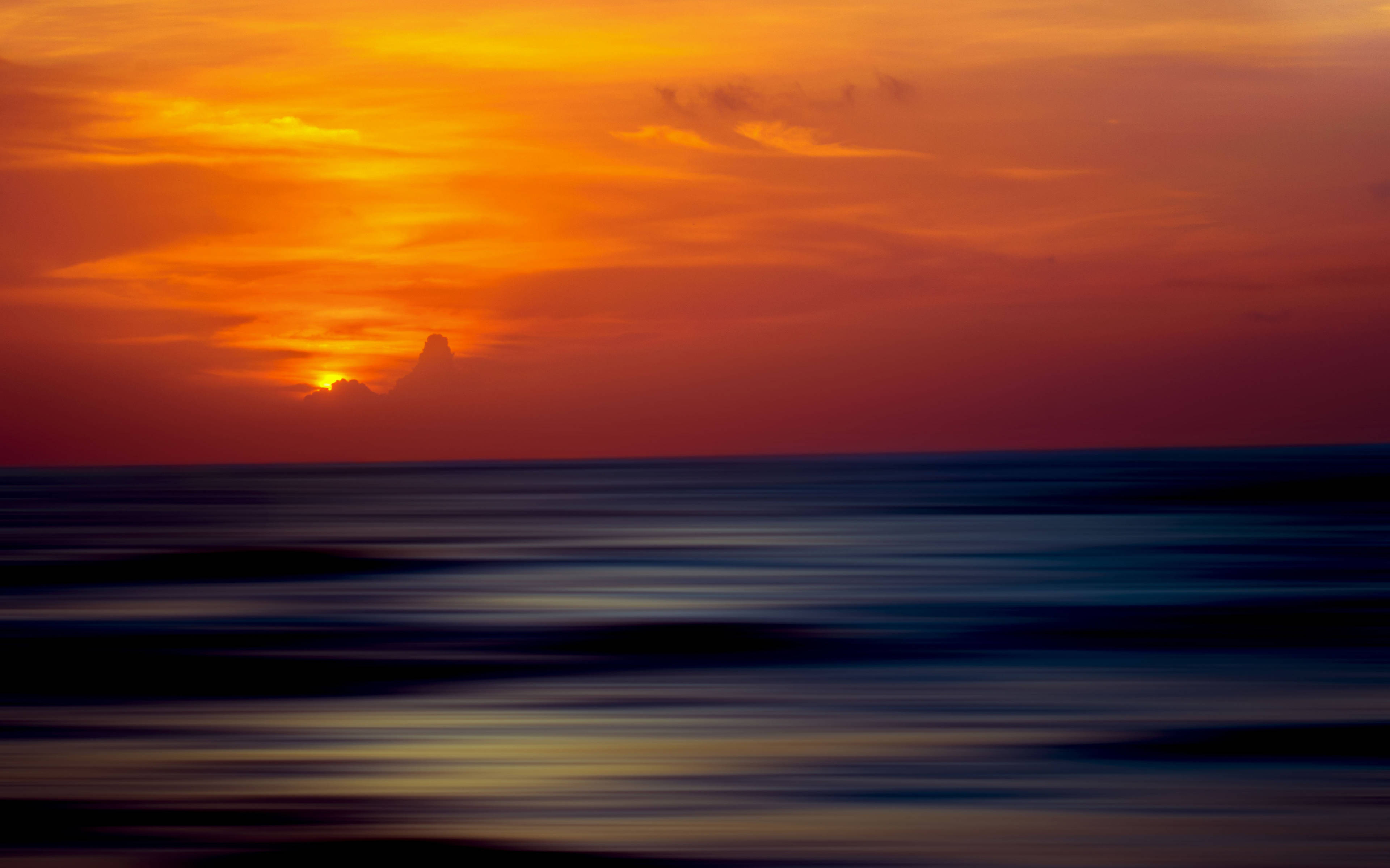 Sunset Beach 桌布 壁紙 — 熾熱的橙色和紅色日落，在動態模糊的海洋地平線上發光，伴隨著孤獨的雲朵剪影。