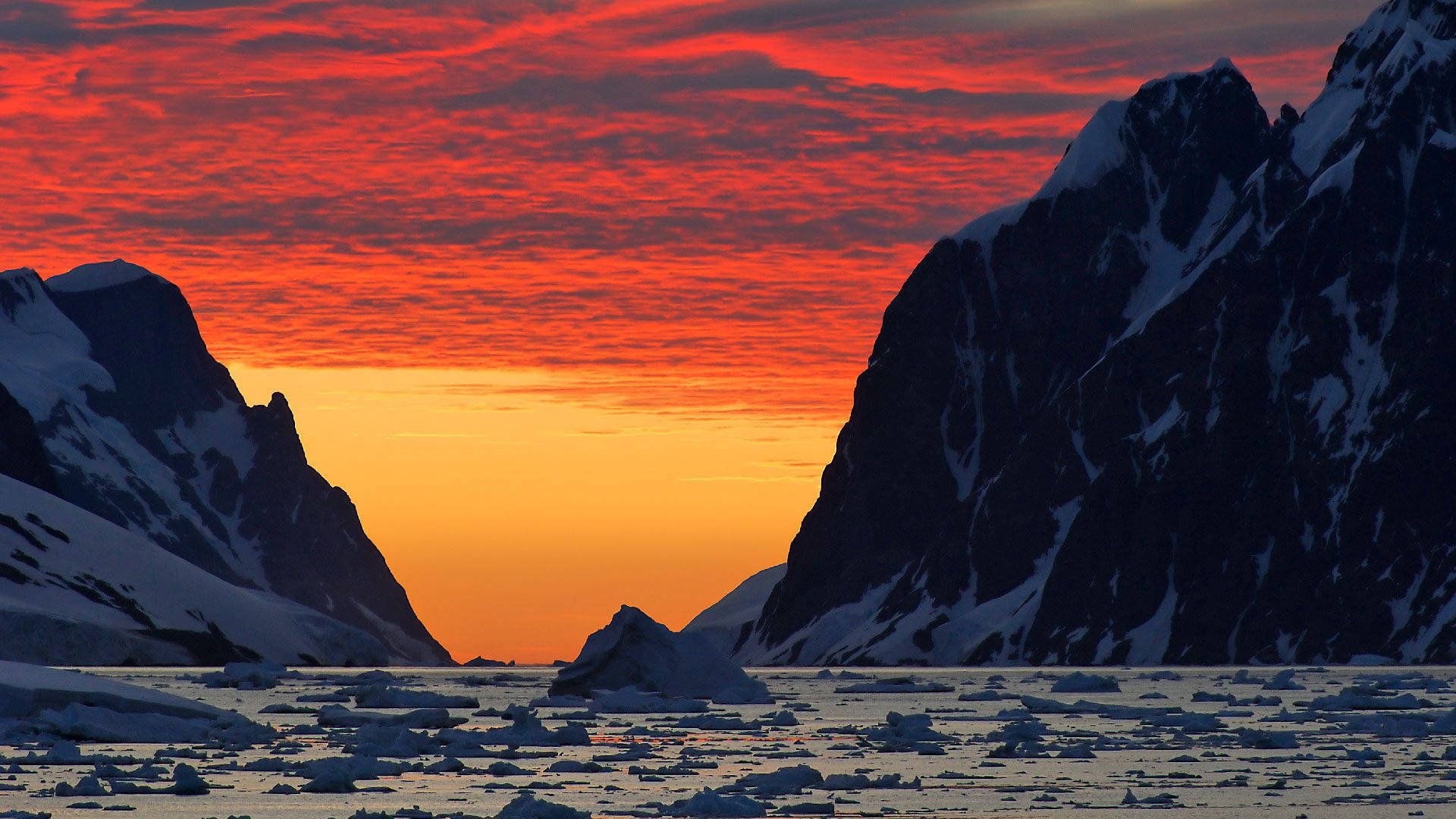Antarctica 桌布 壁紙 — 熾熱的橙色和紅色日落照耀在雪山後面，框住了一片漂浮的冰山海洋。