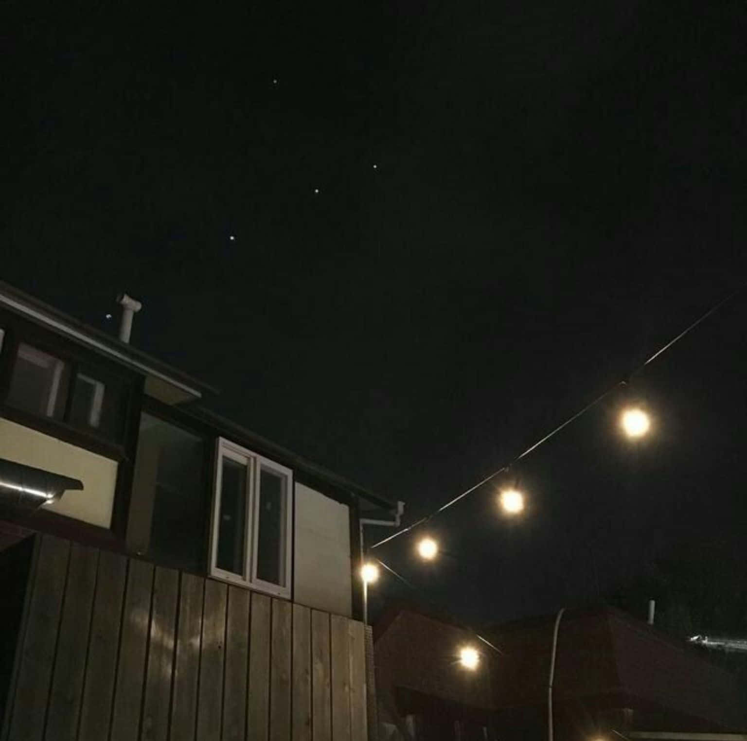 暗黑風格 桌布 壁紙 — 燈串在黑暗的夜空下溫暖地發光，夜空中有可見的星星點綴在木柵欄和房屋上方。