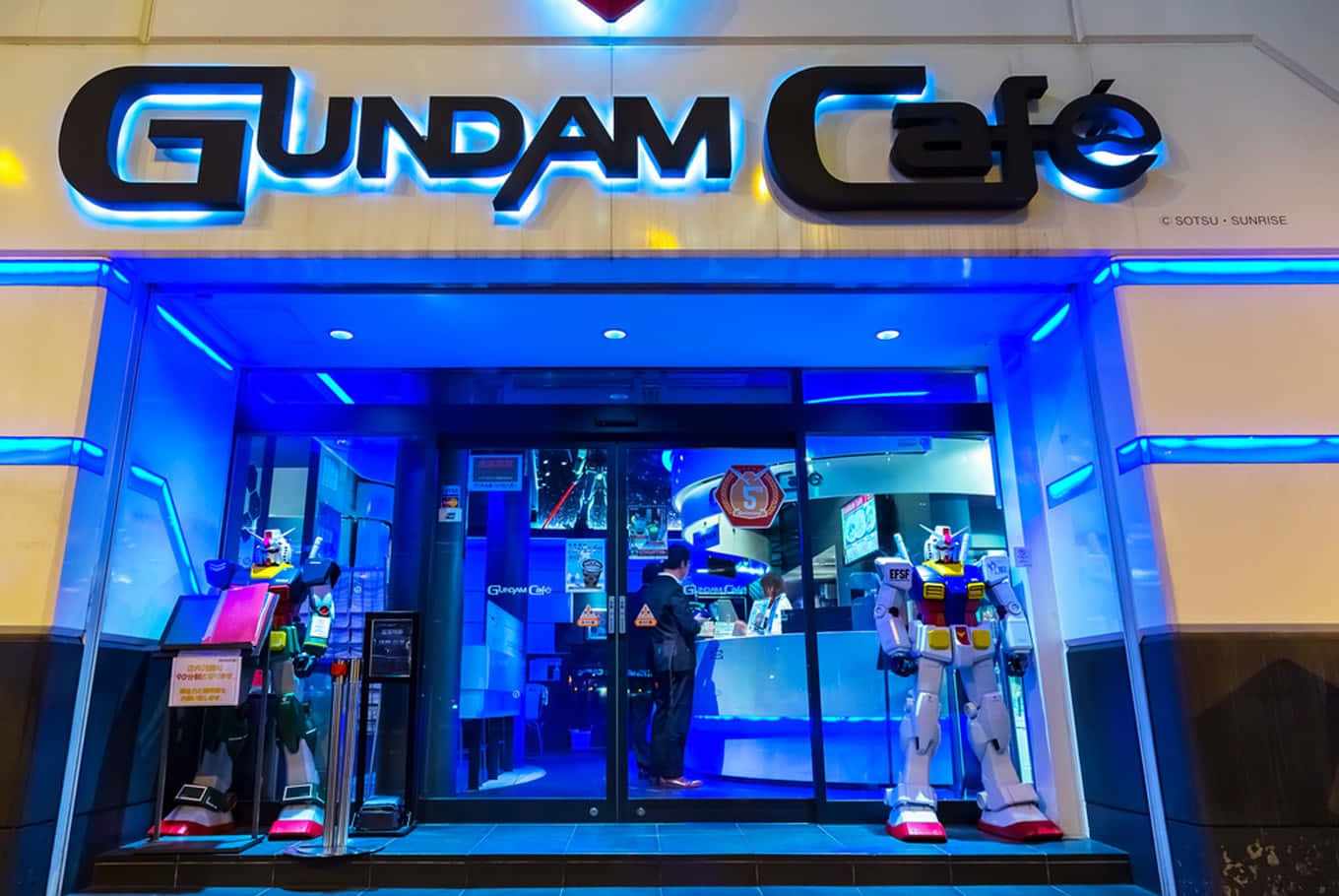Tokyo Japan 桌布 壁紙 — 燈光明亮的Gundam Cafe入口，兩尊大型機器人雕像守衛著玻璃門，上方有發光的藍色霓虹招牌。