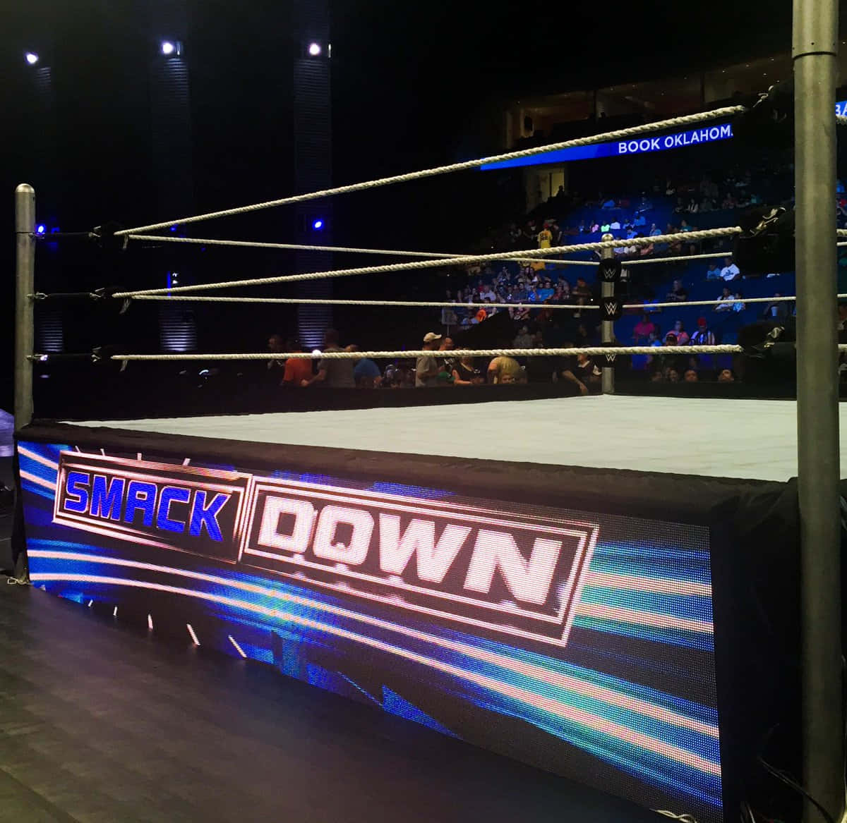 Wrestling Ring 桌布 壁紙 — 燈光明亮的WWE SmackDown摔角擂台，帶有繩索和發光的藍白標誌，周圍是黑暗的競技場，看台上有觀眾。