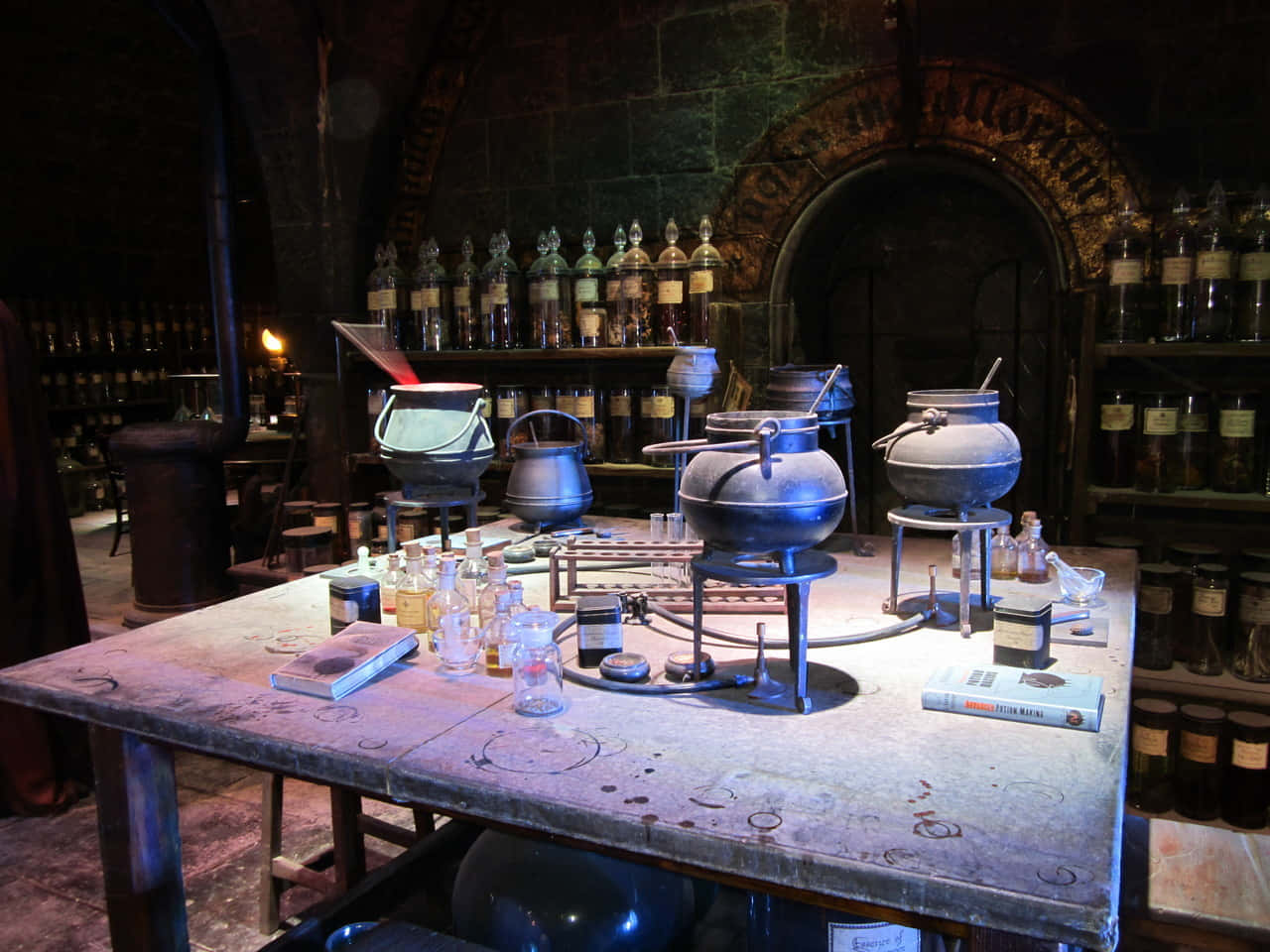 The Hogwarts Dark Arts Classroom 桌布 壁紙 — 燈光昏暗的中世紀風格藥水實驗室，有冒泡的大鍋和裝滿發光液體的玻璃瓶架。