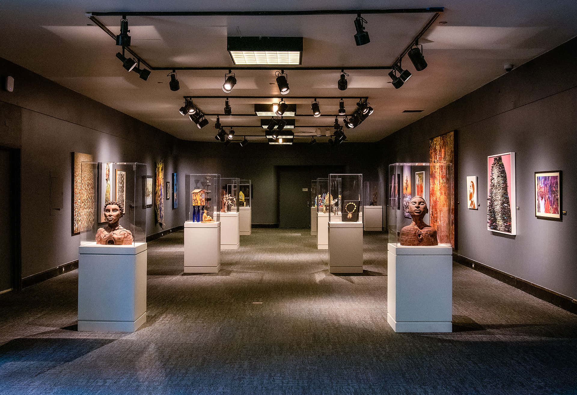 Exhibition 桌布 壁紙 — 燈光昏暗的博物館畫廊，聚光燈照亮玻璃櫃中的半身像和灰色牆壁上色彩繽紛的畫作。