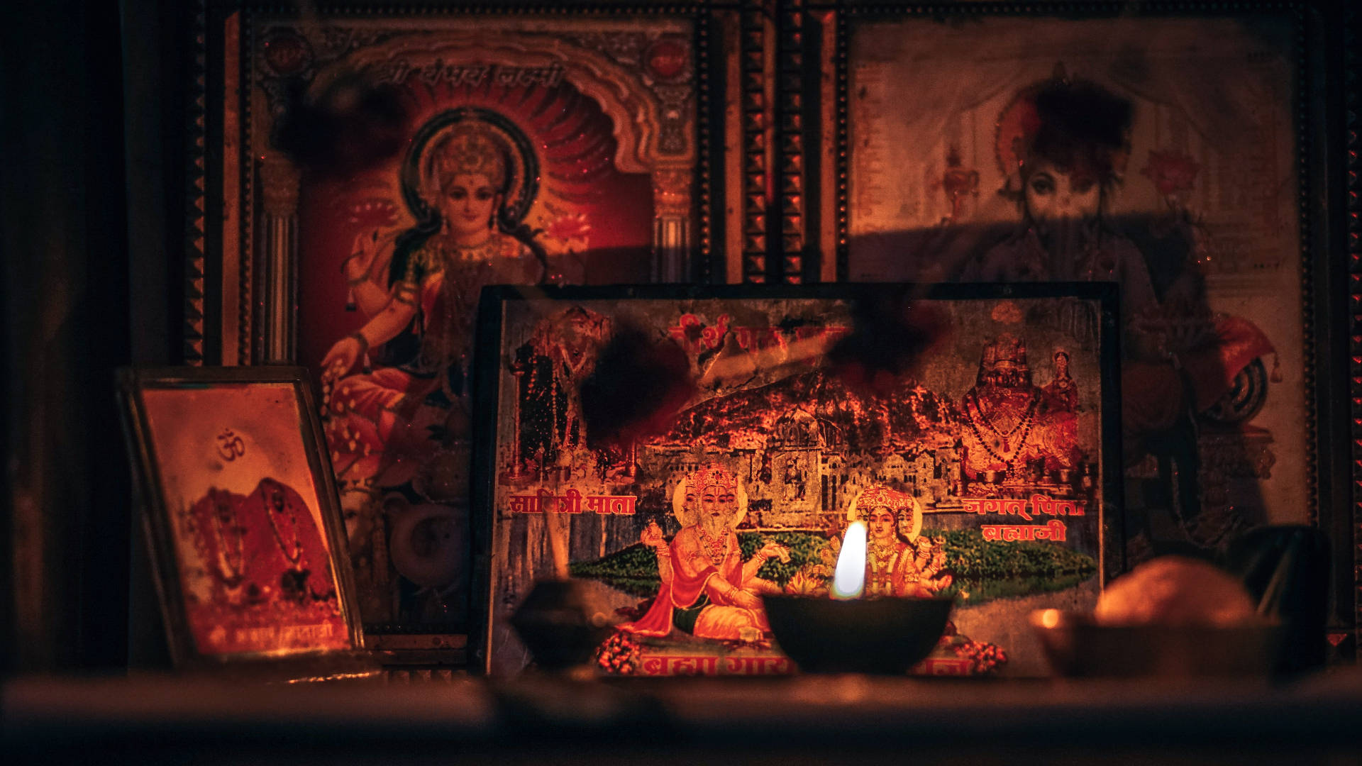 Hindu God 桌布 壁紙 — 燈光昏暗的印度教神龕，帶有框裝的神像和一支發光的蠟燭，將溫暖的光線投射在神聖的圖標上。
