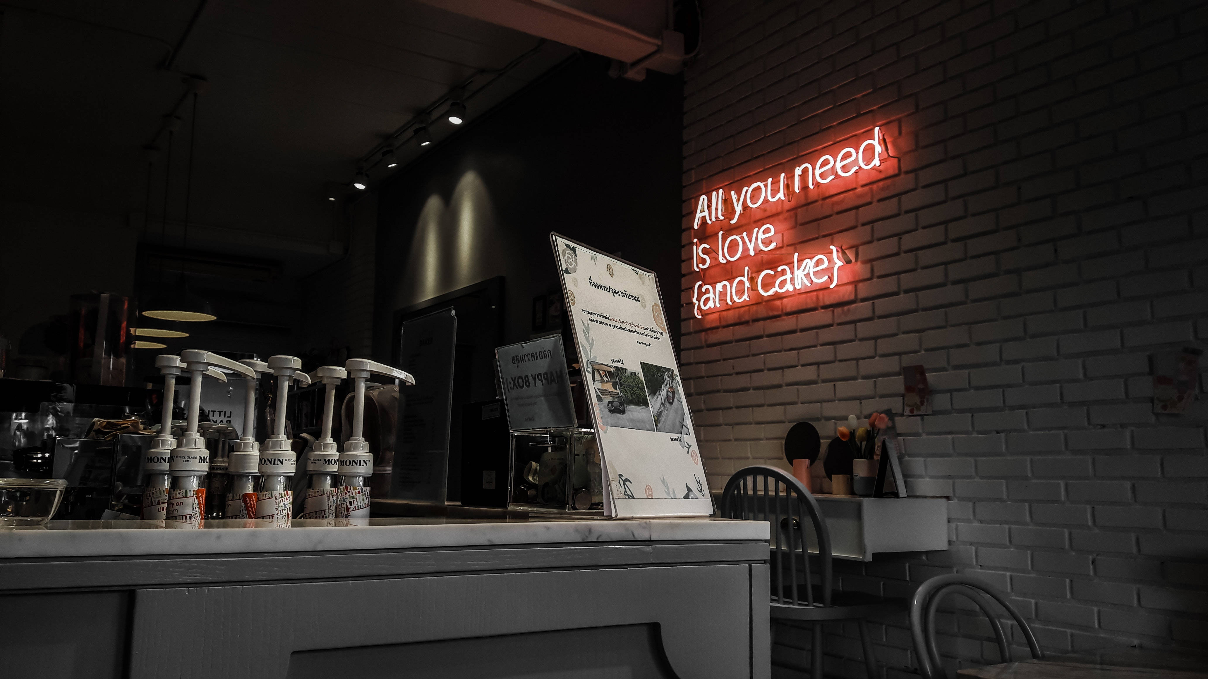 neon sign 桌布 壁紙 — 燈光昏暗的咖啡館，白色磚牆上有一個發光的紅色霓虹燈牌，上面寫著「你所需要的只是愛{和蛋糕}」。