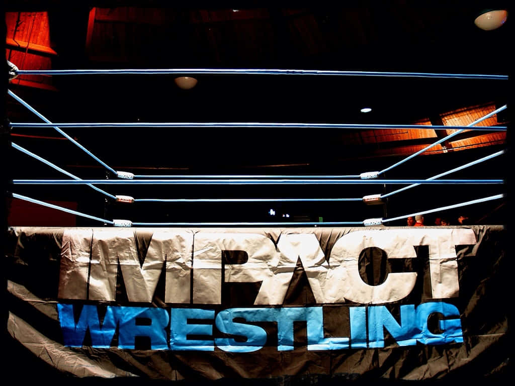 Wrestling Ring 桌布 壁紙 — 燈光昏暗的摔角擂台，藍色繩索和一個皺巴巴的橫幅，上面寫著「IMPACT WRESTLING」，在戲劇性的頂燈照射下。