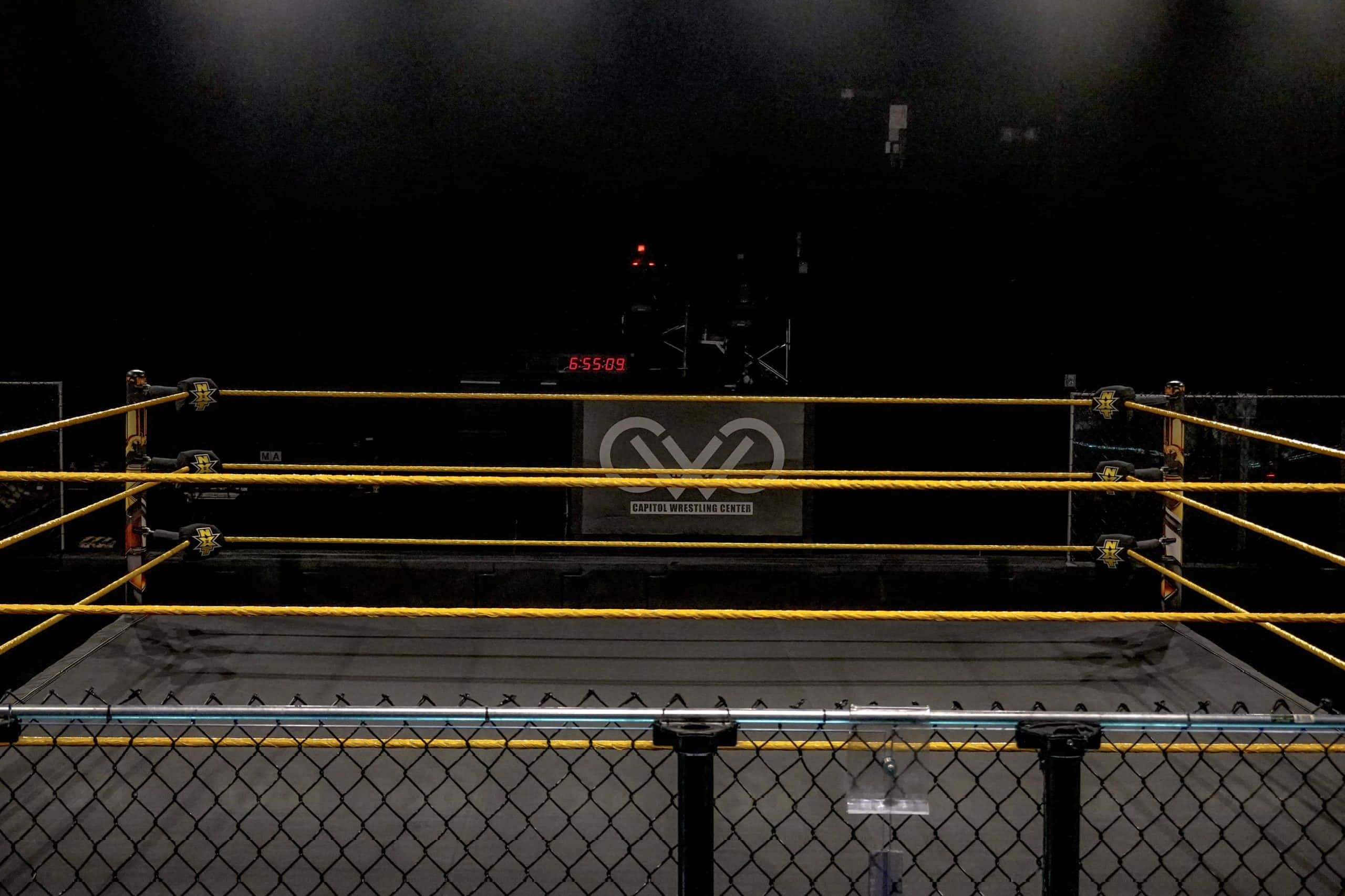 Wrestling Ring 桌布 壁紙 — 燈光昏暗的摔角擂台，黃色繩索和數位時鐘顯示6:55:09，在聚光燈下。
