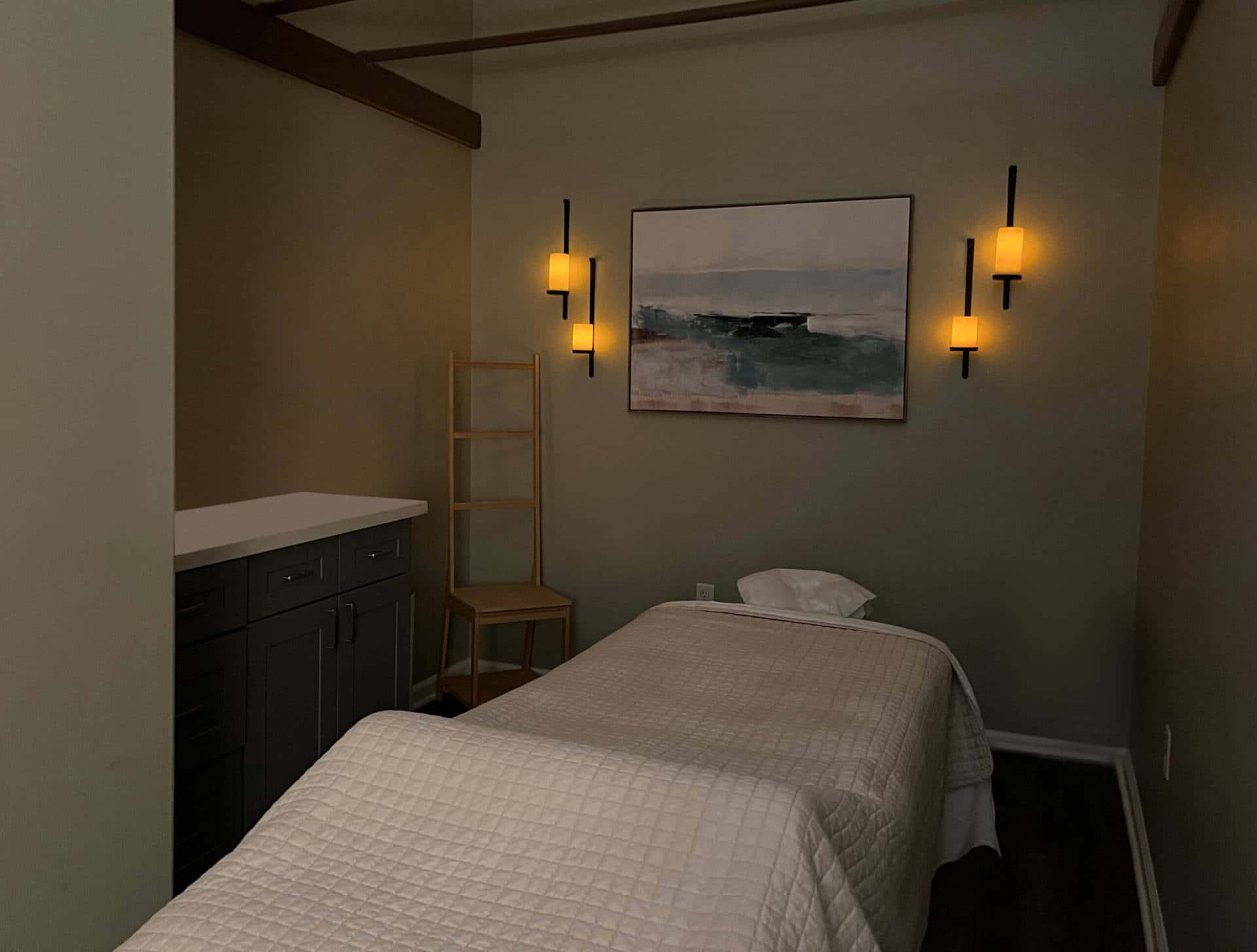 Massage Room 桌布 壁紙 — 燈光昏暗的水療室，設有按摩床，暖色壁燈下掛著寧靜的海洋畫作。