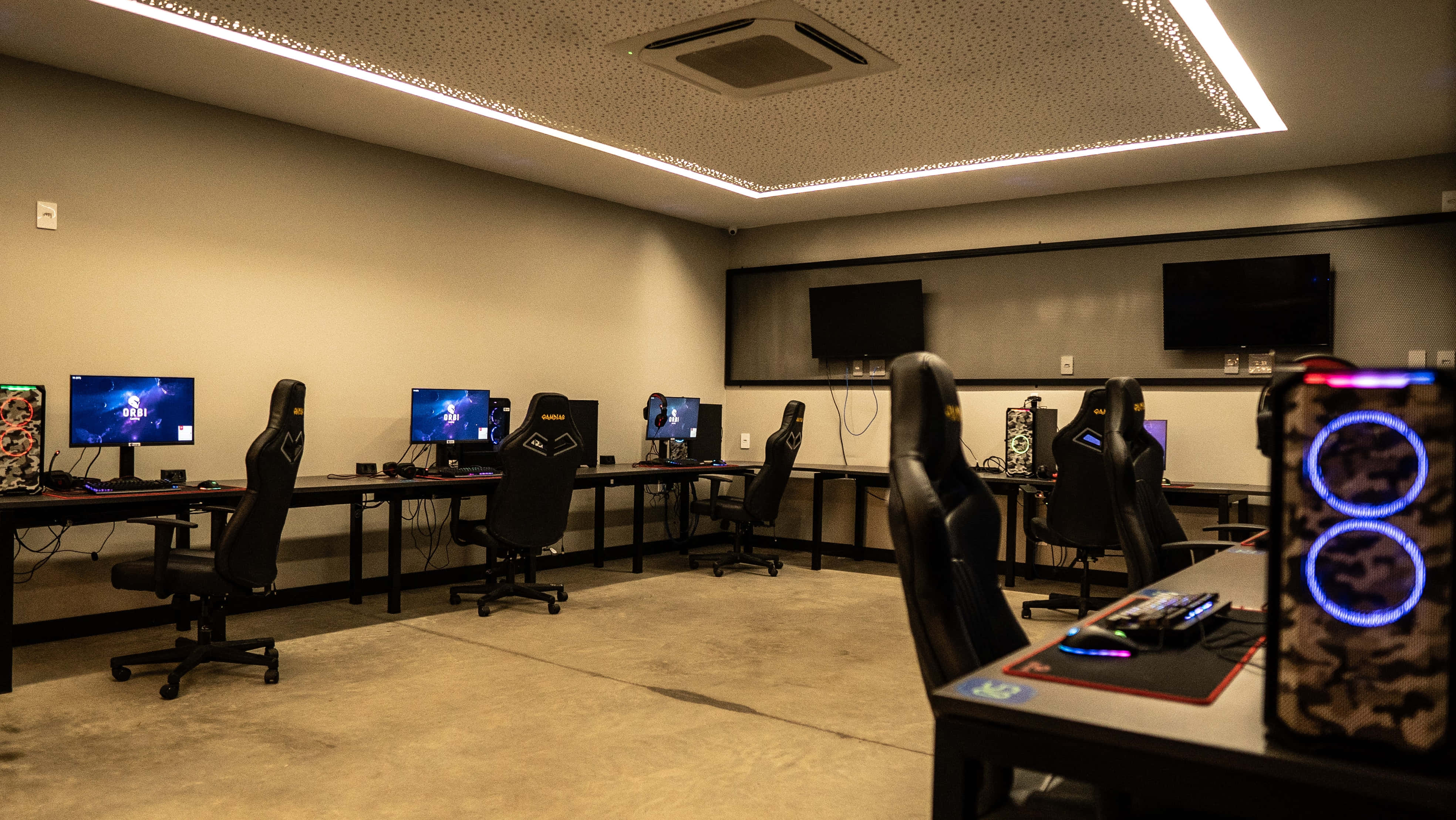 Gaming Room 桌布 壁紙 — 燈光昏暗的遊戲休息室，現代天花板燈光下排列著黑色人體工學椅和發光的電腦設置。