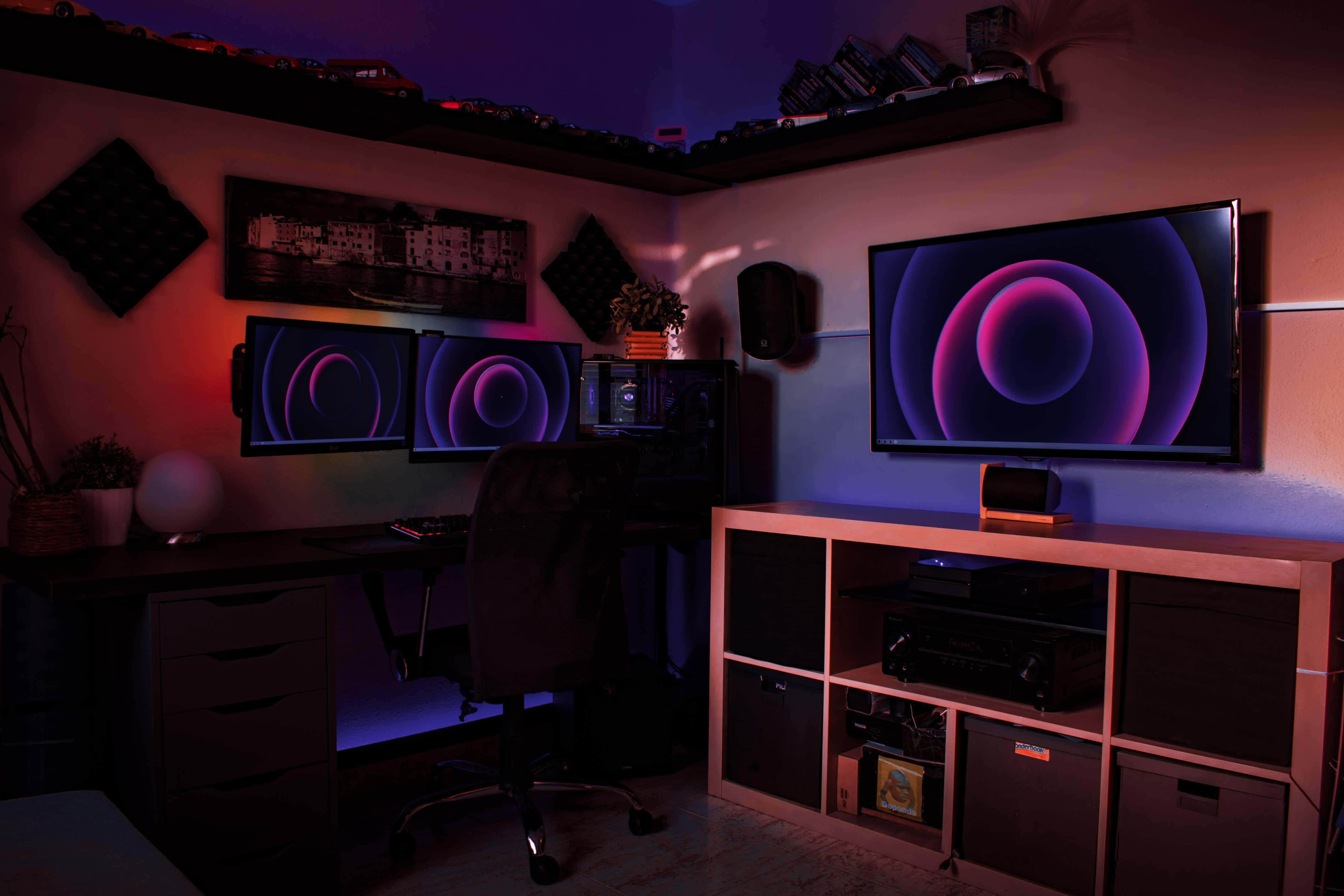 Gaming Room 桌布 壁紙 — 燈光昏暗的遊戲室，多個顯示器顯示抽象圓形圖案，沐浴在紫色和橙色的光芒中。