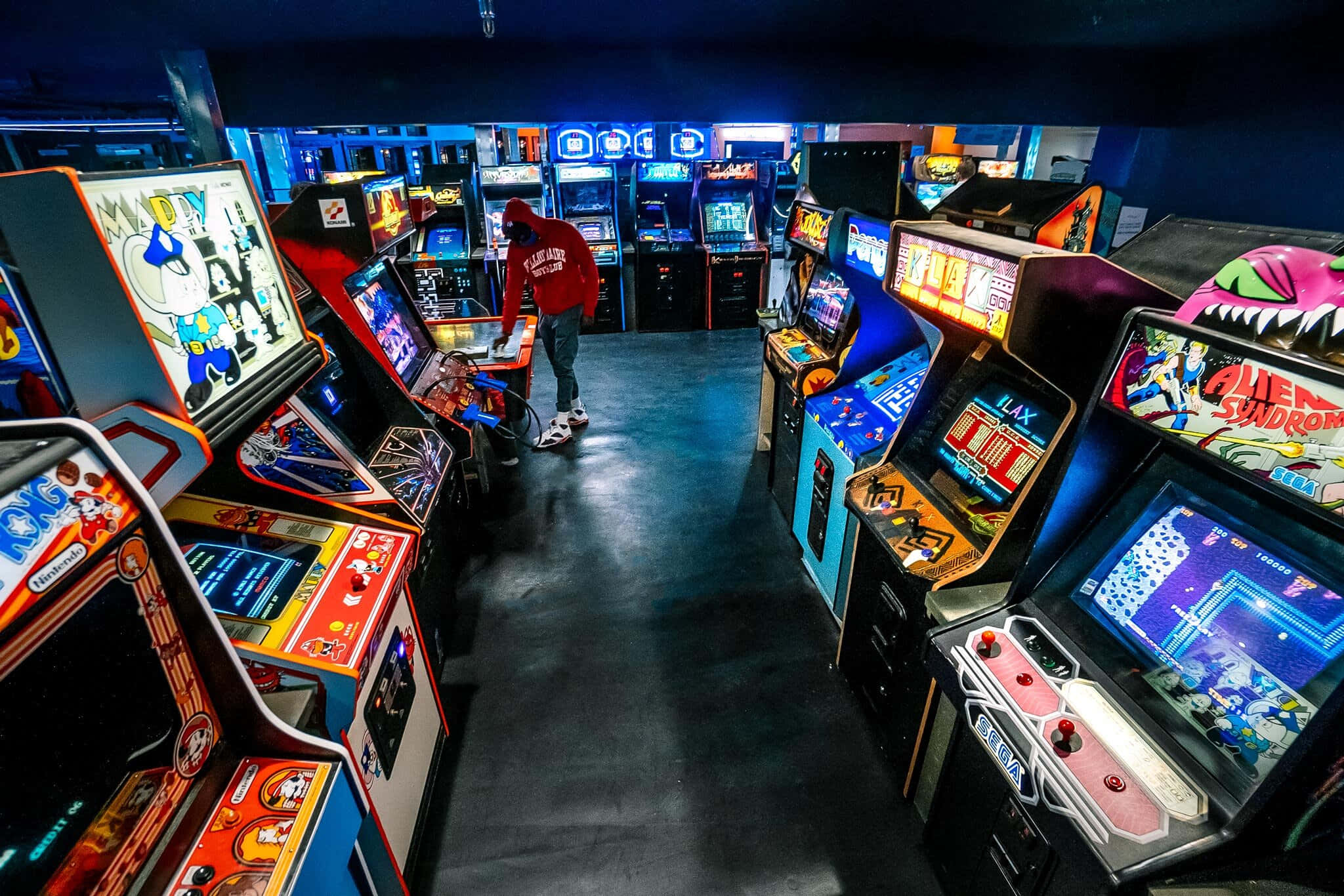Arcade Games 桌布 壁紙 — 燈光昏暗的遊戲機中心，充滿發光的復古遊戲機，中間有一人在玩。