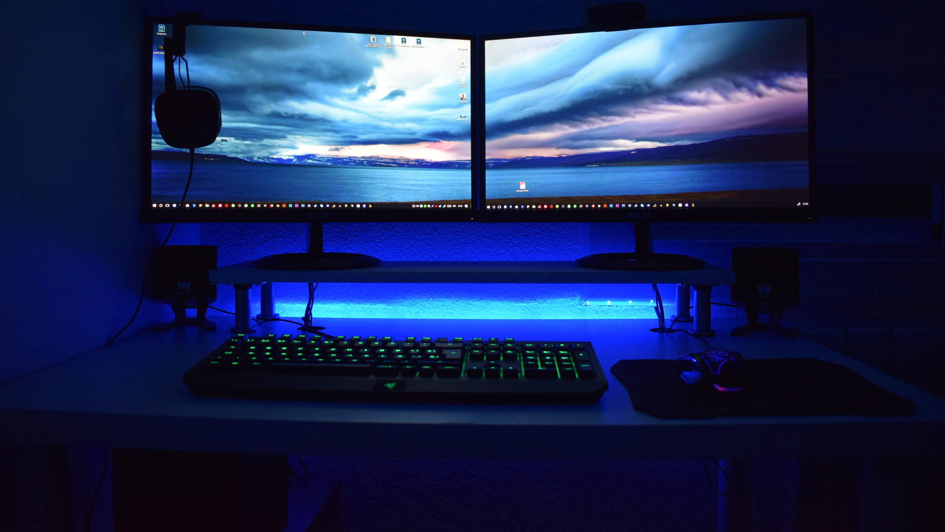 Desk 桌布 壁紙 — 燈光昏暗的遊戲設置，雙顯示器顯示水上暴風雨日落，綠色發光鍵盤在藍色環境光下。