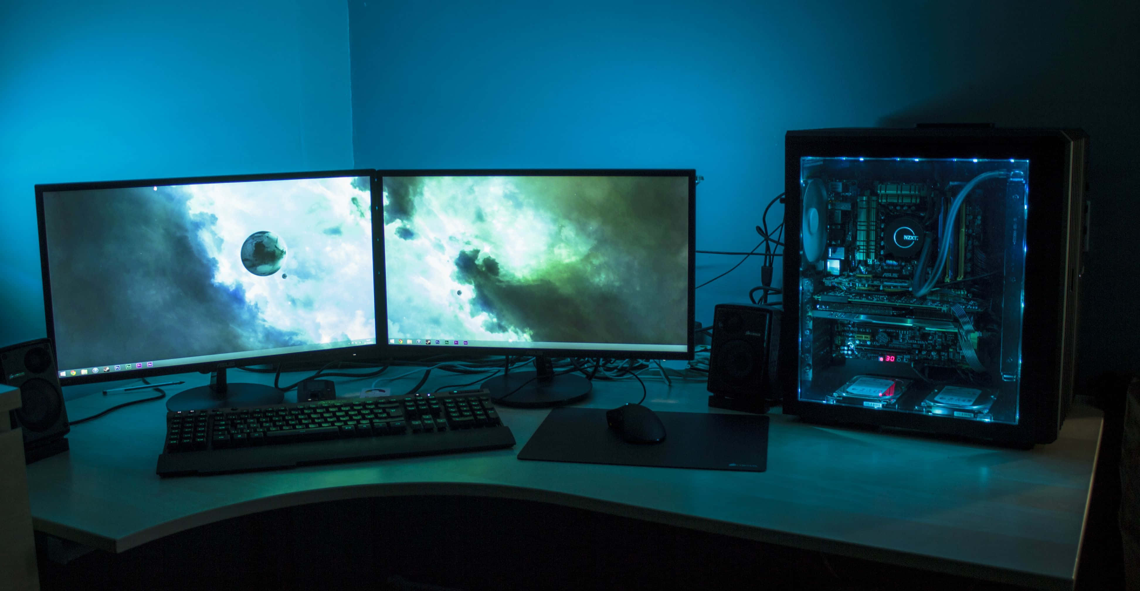 Gaming Setup 桌布 壁紙 — 燈光昏暗的遊戲設置，雙顯示器顯示宇宙場景，桌上有一個發光的透明PC機箱。