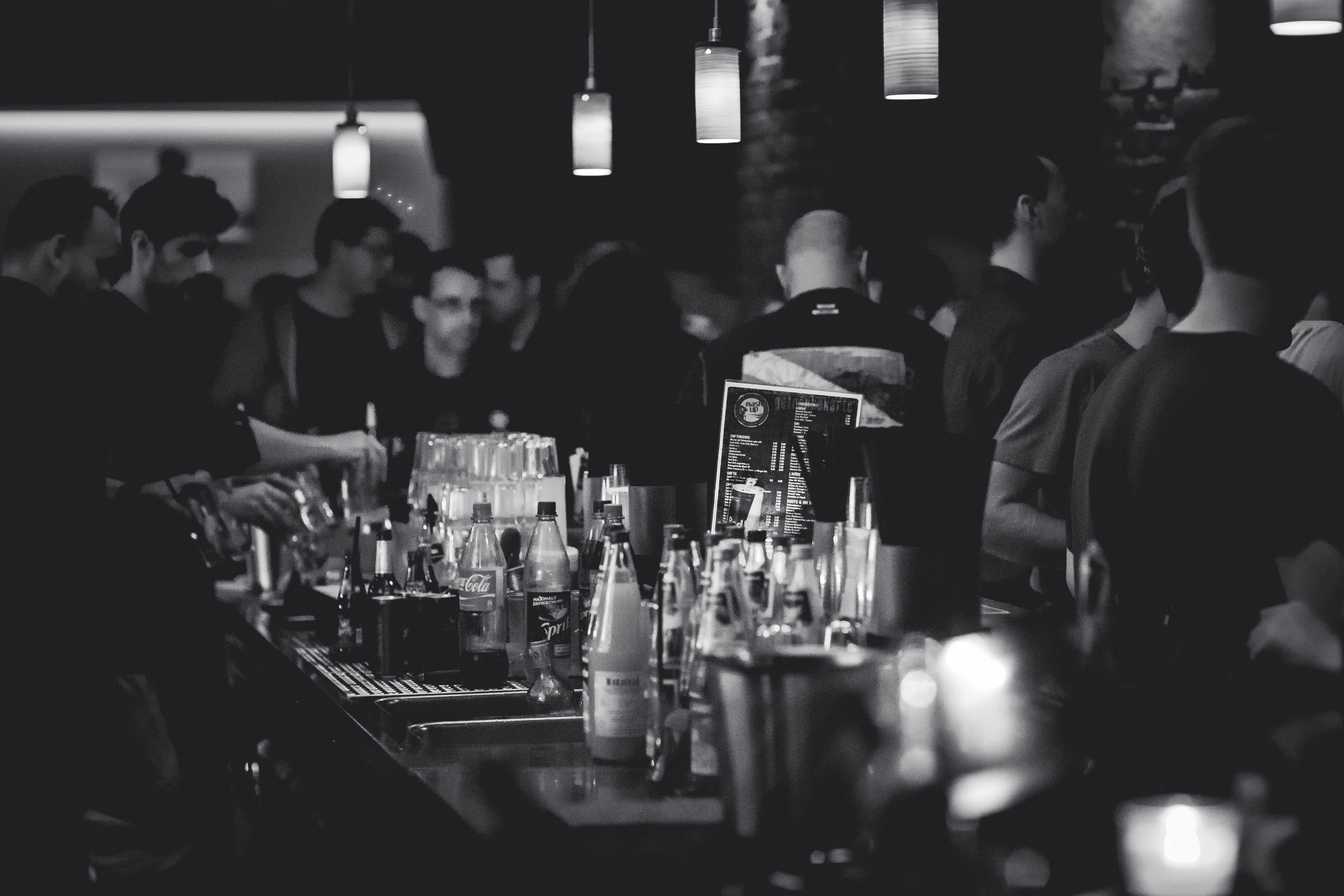 Bar 桌布 壁紙 — 燈光昏暗的酒吧，顧客聚集在吧檯周圍，調酒師在吊燈下調製飲品。