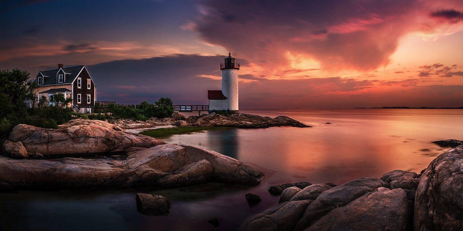 Lighthouse 桌布 壁紙 — 燈塔和海岸房屋在熾熱的日落天空下閃耀，倒映在崎嶇岩石環繞的平靜水域中。