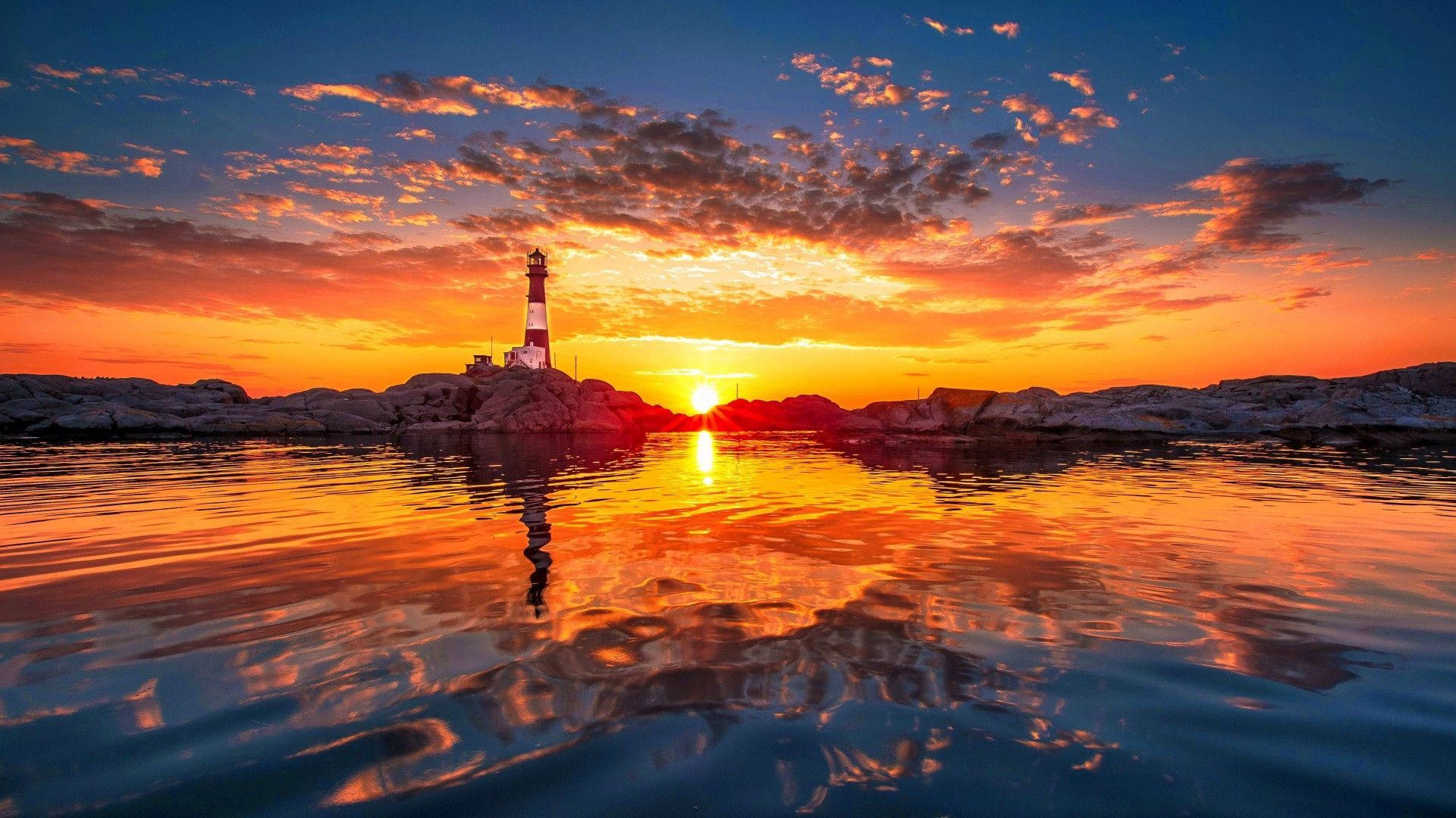 Ocean Sunset 桌布 壁紙 — 燈塔矗立在岩石海岸上，夕陽西下，在平靜的水面上投射出火熱的橙色和粉紅色倒影。