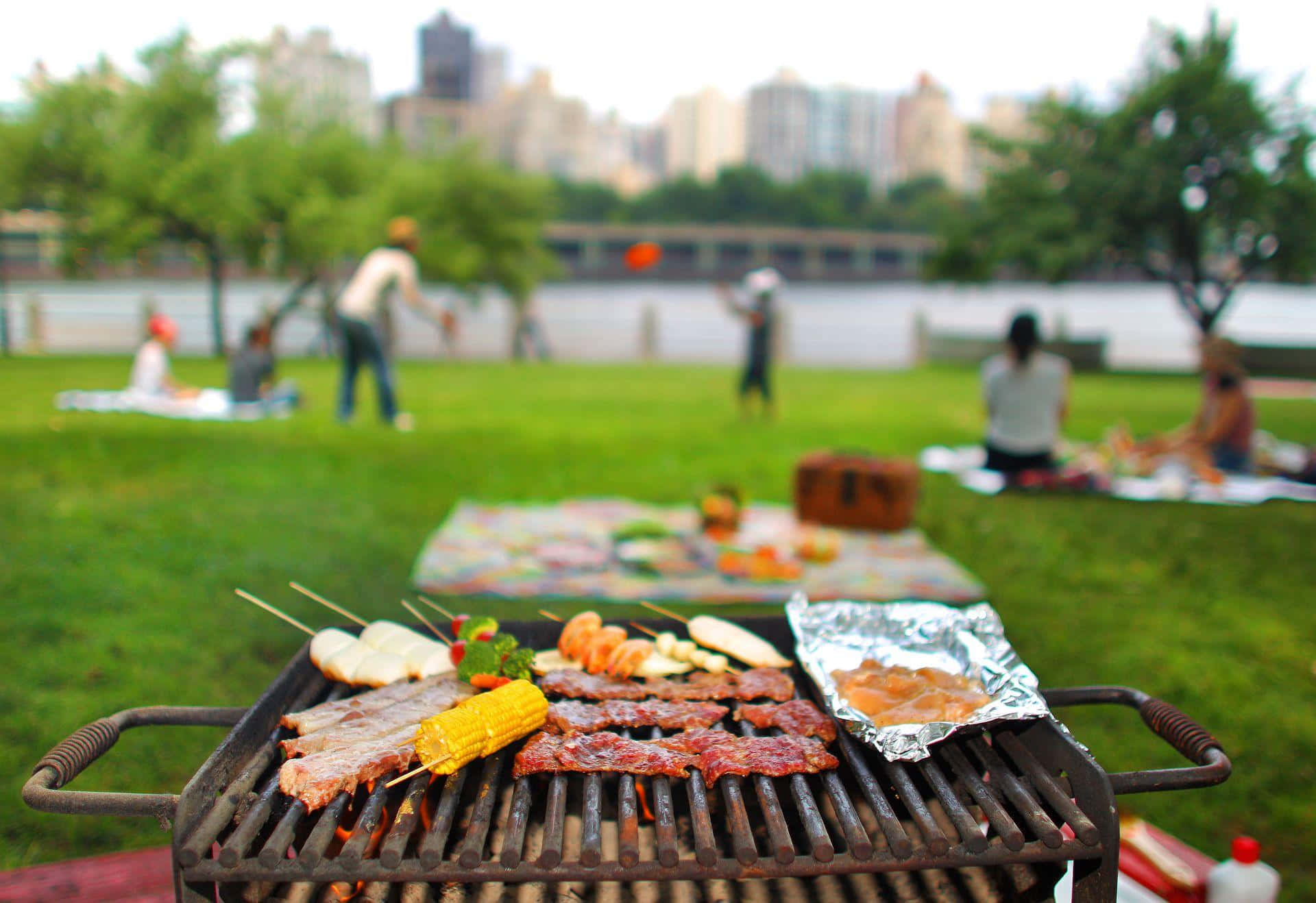 picnic 桌布 壁紙 — 燒烤架上滋滋作響的烤肉和玉米特寫，背景是公園裡野餐的人們。