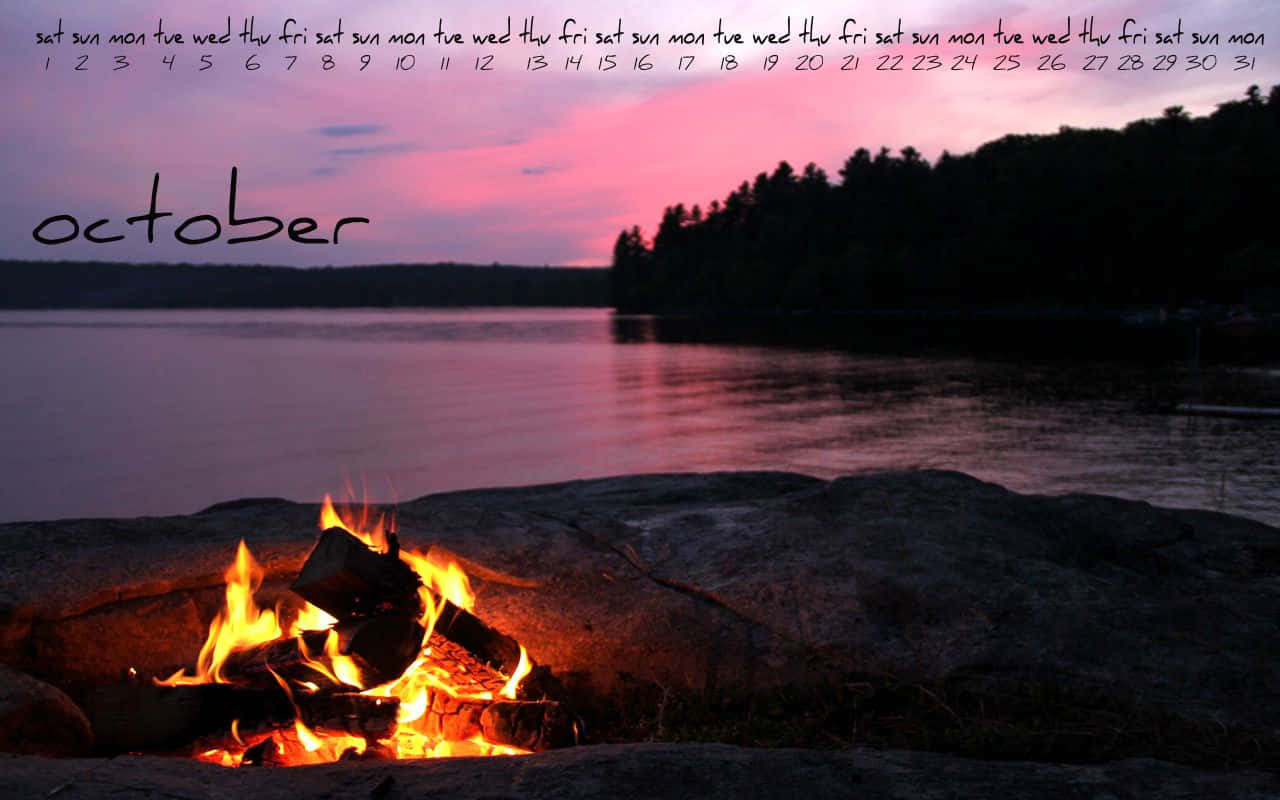 Campfire 桌布 壁紙 — 營火在岩石海岸上劈啪作響，粉紅色的日落映照在樹林山丘後方的平靜水面上。