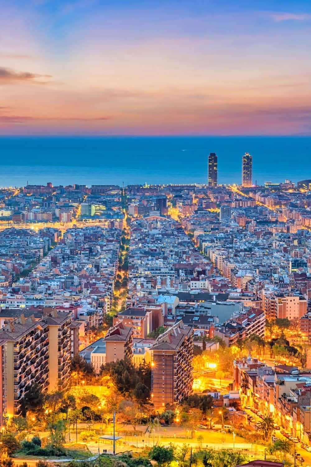 Barcelona 桌布 壁紙 — 燦爛的夕陽以溫暖的色調沐浴著廣闊的海濱城市景觀，燈光在建築物和街道間閃爍。