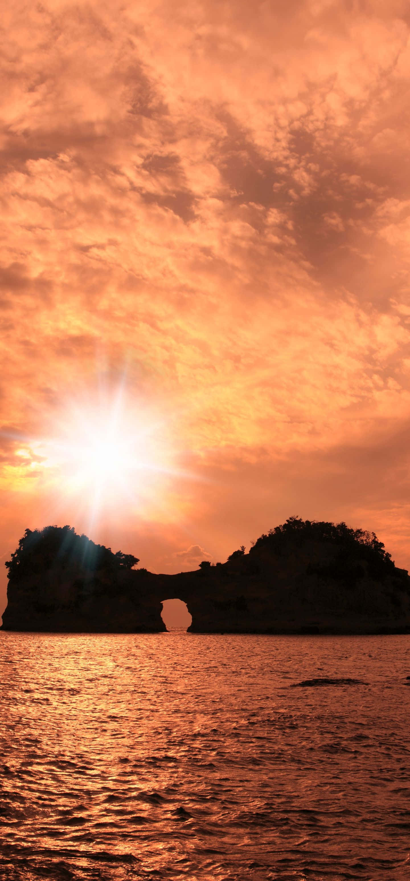 1440x3088 桌布 壁紙 — 燦爛的夕陽在平靜的海面上投射出熾熱的橙色光芒，在戲劇性的天空下照亮了天然岩石拱門的輪廓。