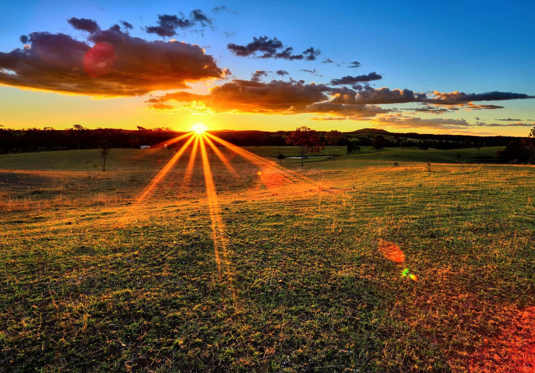 太陽 桌布 壁紙 — 燦爛的夕陽在戲劇性的天空下，將金色光芒灑落在廣闊的草地上，天空中有散落的雲朵。