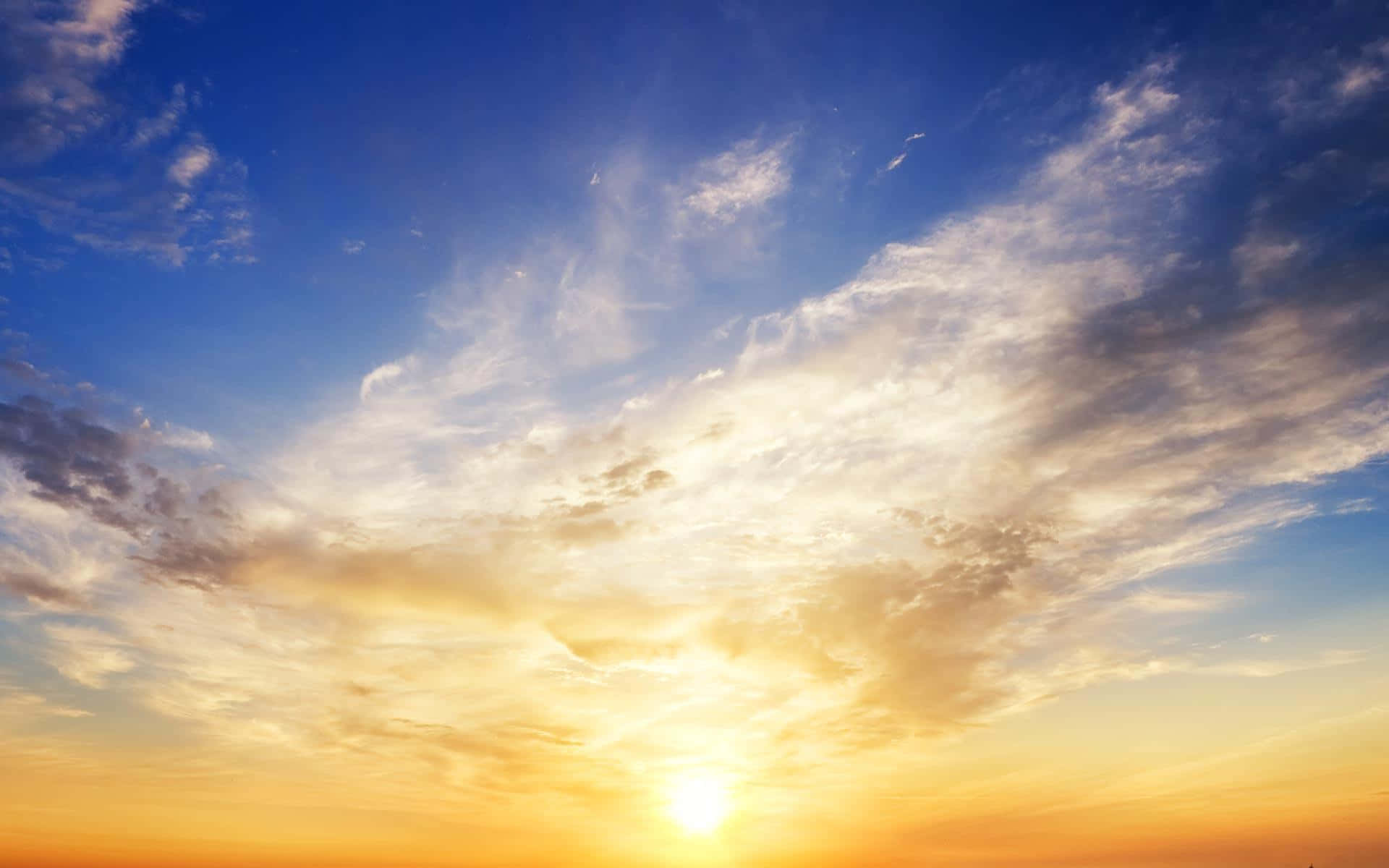 Sky Clouds 桌布 壁紙 — 燦爛的夕陽將天空染成金橙色和深藍色，薄雲捕捉著太陽的光芒。