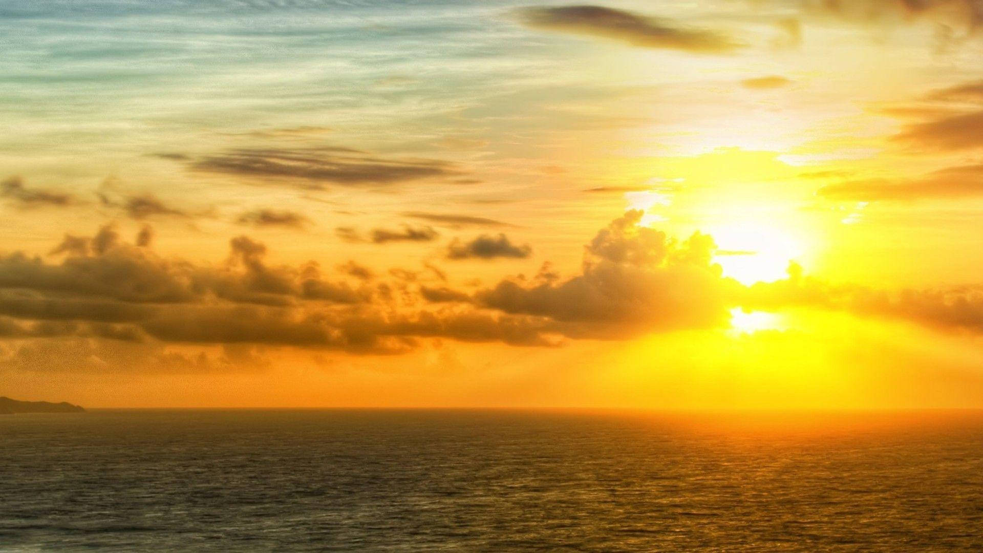 Sunset Sky 桌布 壁紙 — 燦爛的夕陽將海洋染成金色，雲朵在柔和的藍天下閃耀琥珀色光芒。