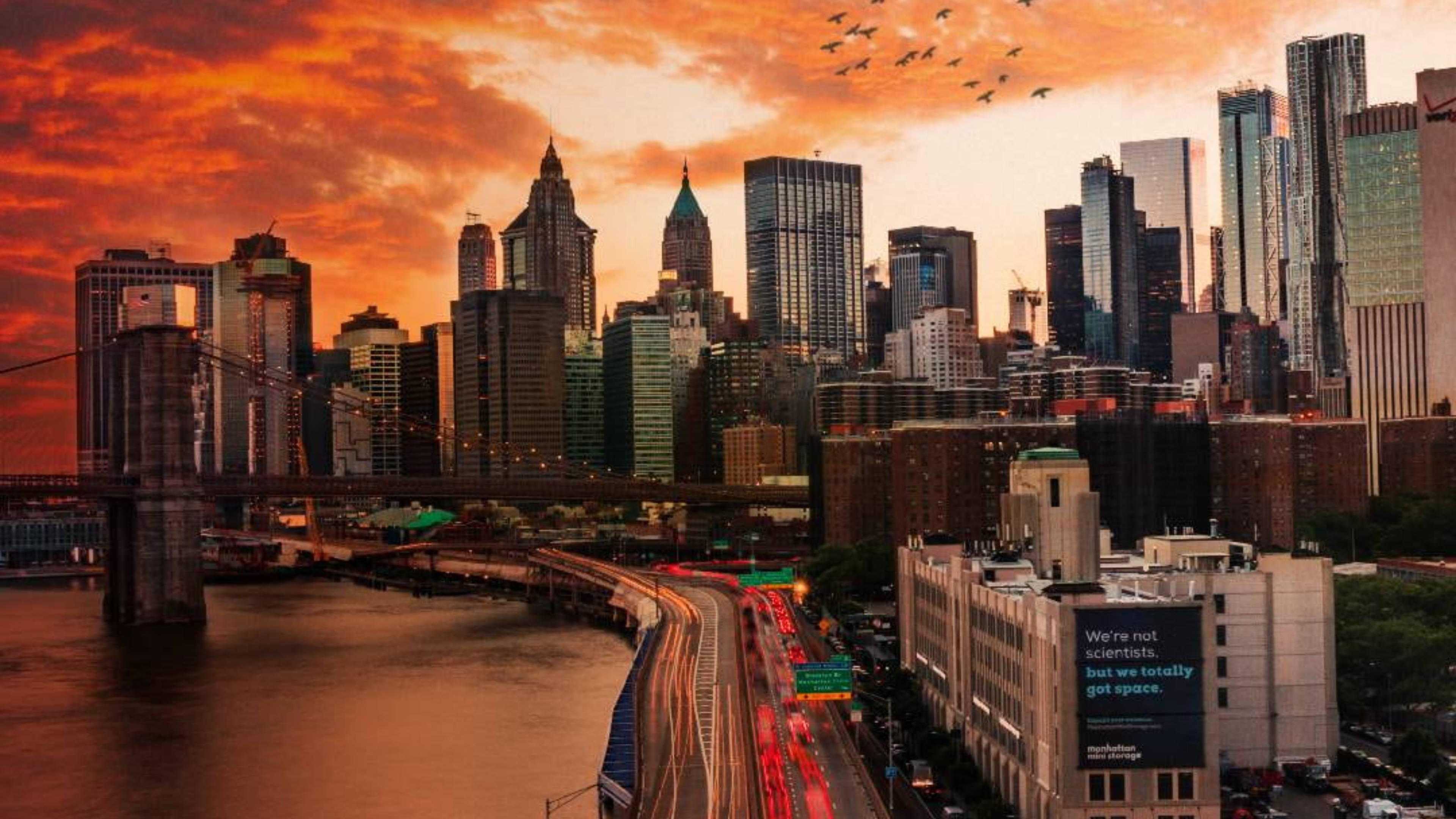 New York 桌布 壁紙 — 燦爛的夕陽將紐約市的天際線染成火熱的橙色，橋下的河濱高速公路上車流如織。