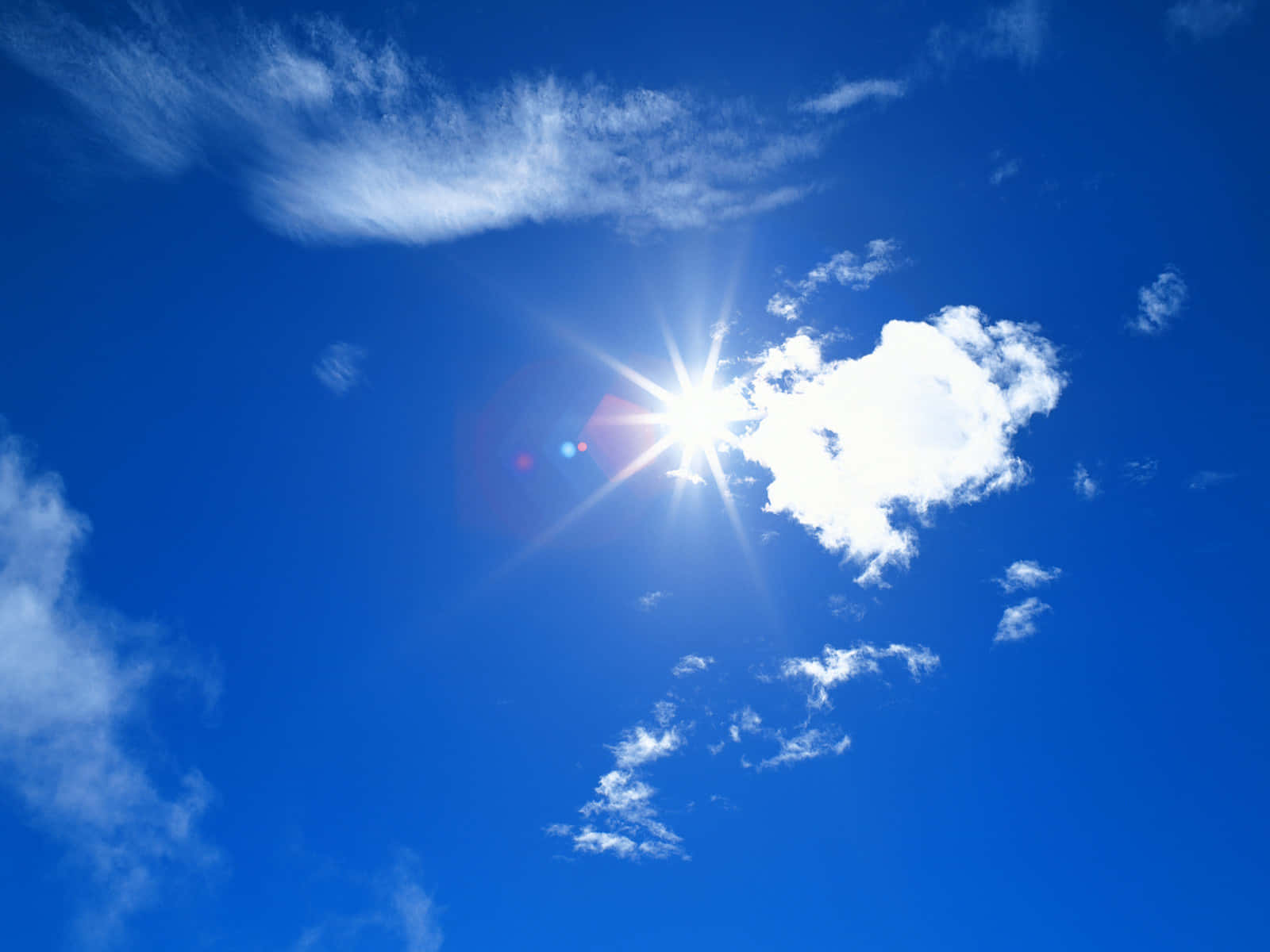 Blue Skies 桌布 壁紙 — 燦爛的太陽在蔚藍的天空中，透過散佈的白雲散發出明亮的光芒。