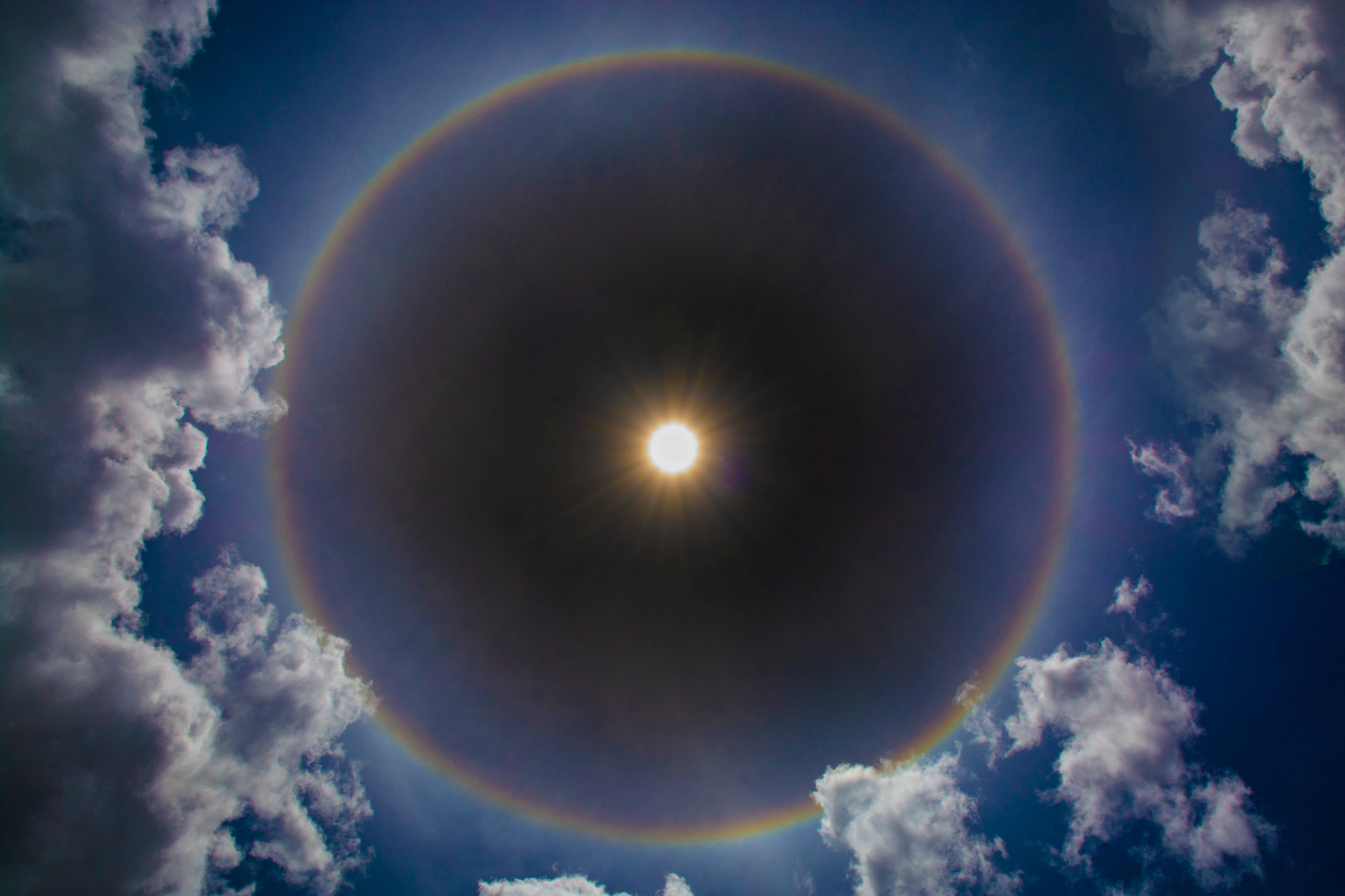 4K 太空 桌布 壁紙 — 燦爛的太陽被鮮豔的彩虹光環環繞，背景是深藍色的天空和蓬鬆的白雲。