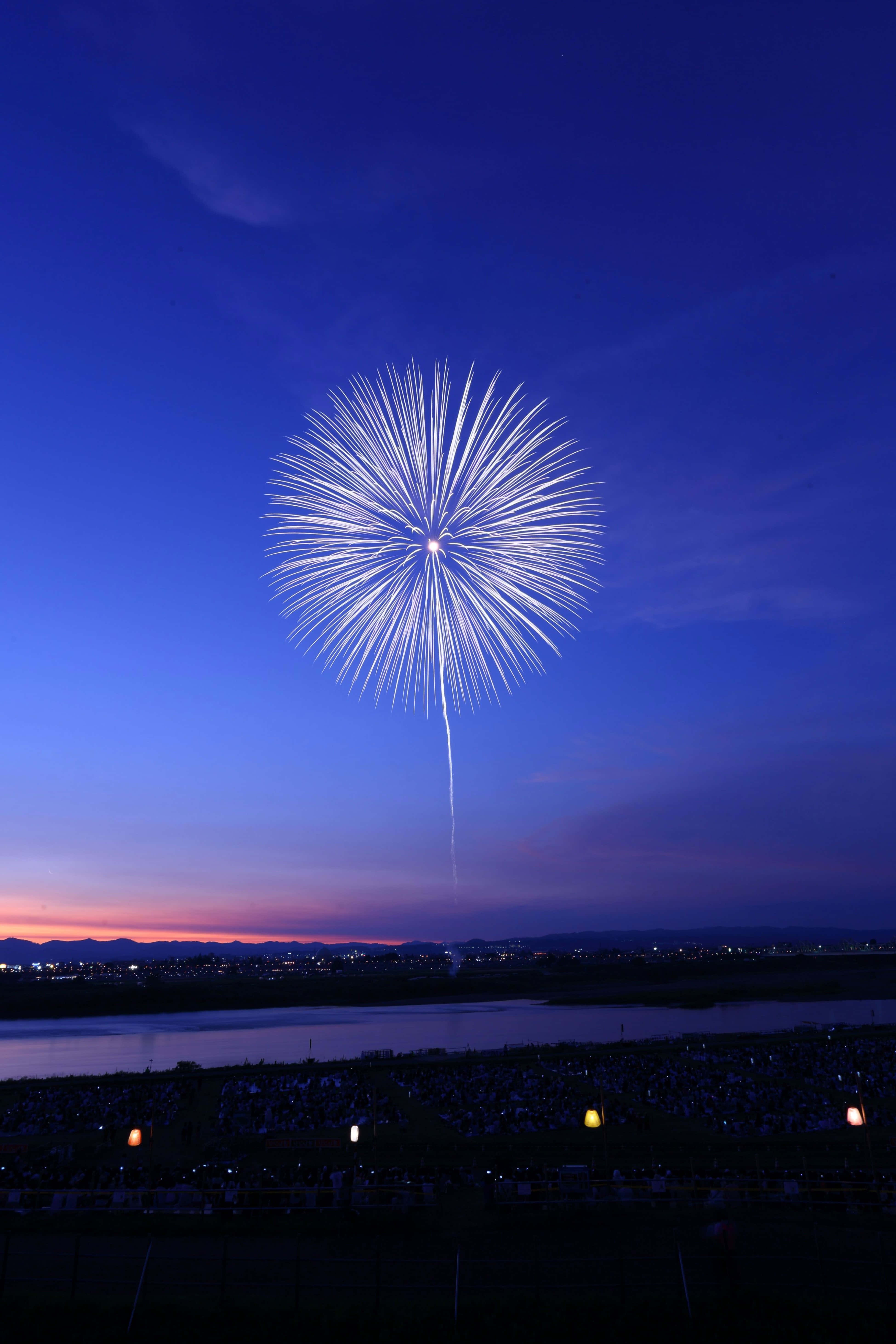 firework 桌布 壁紙 — 燦爛的白色煙火在暮色天空中爆炸，下方是河流和城市景觀，燈籠閃爍。
