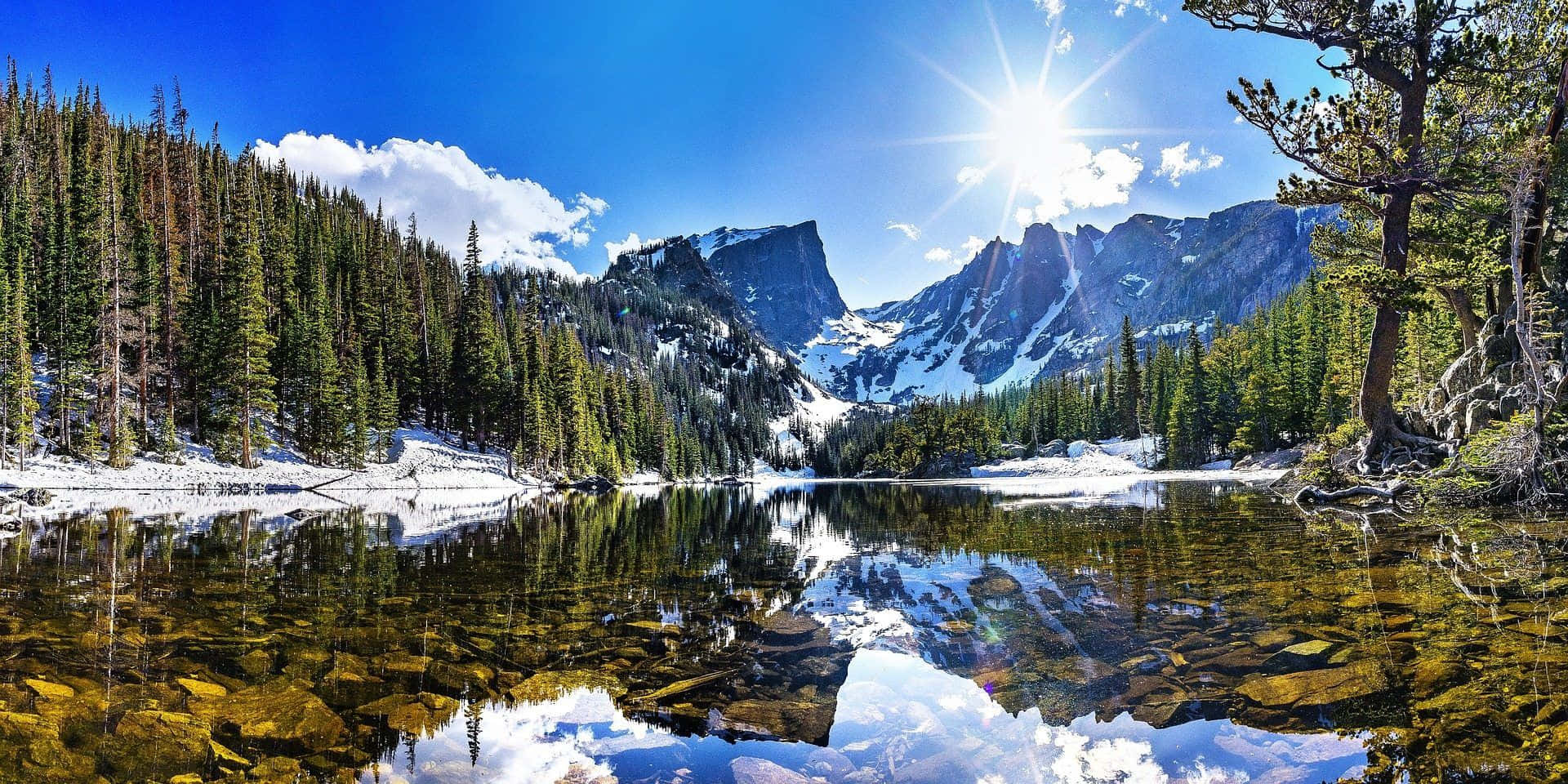 Colorado Mountain 桌布 壁紙 — 燦爛的陽光照耀在積雪的山峰上，完美地倒映在寧靜的高山湖泊中，周圍是常綠森林。