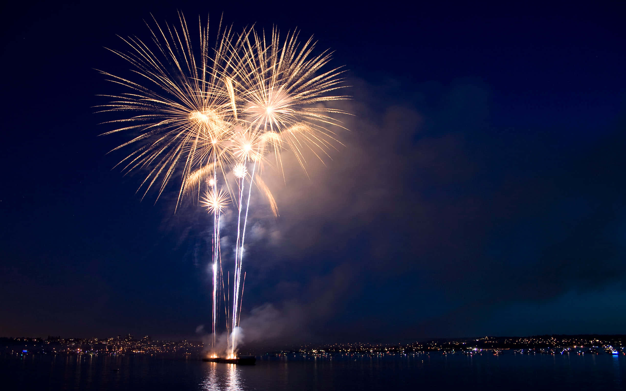 Fireworks 桌布 壁紙 — 爆炸性的金色煙火在黑暗的水面上綻放，遠岸的城市燈光閃爍。