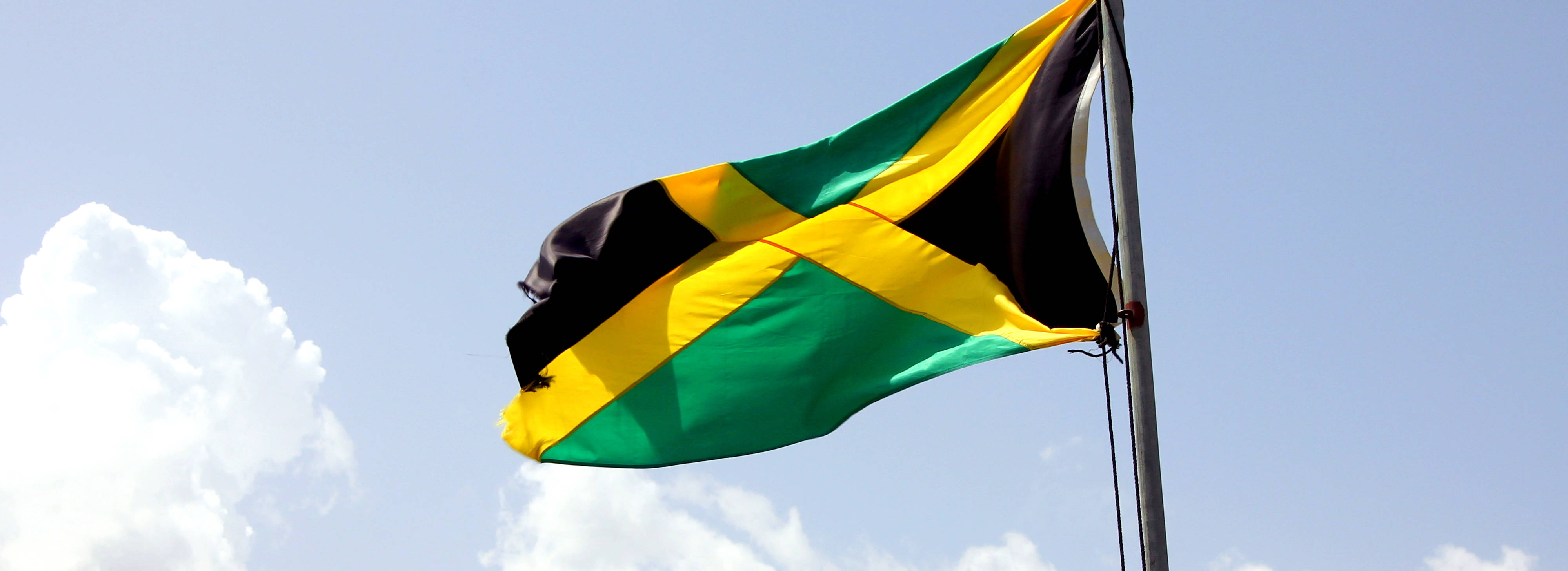 Jamaica 桌布 壁紙 — 牙買加國旗，帶有綠色、黃色和黑色條紋，在晴朗的藍天和蓬鬆的白雲中勇敢飄揚。