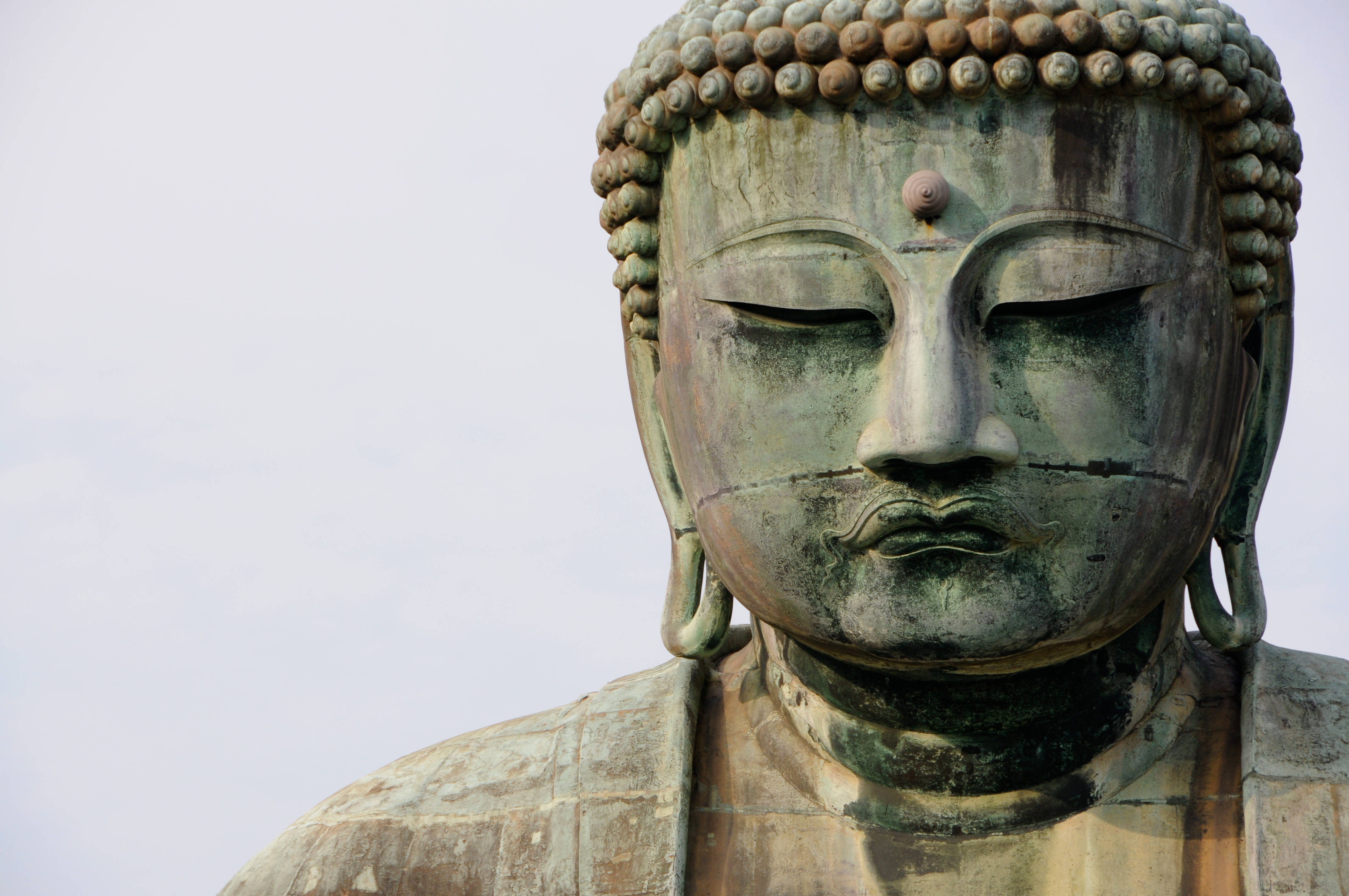 禪 桌布 壁紙 — 特寫：一尊寧靜、風化的青銅佛像，閉著眼睛，帶有精細的肉髻，背景是淡色天空。