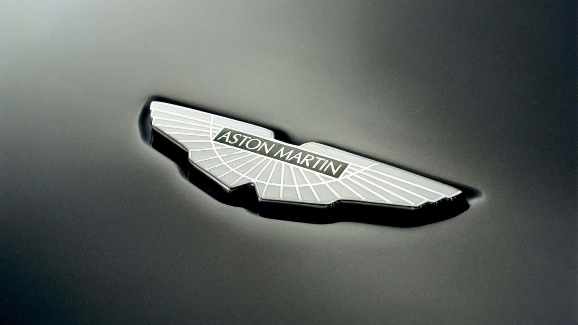 奧斯頓馬丁 桌布 壁紙 — 特寫：銀色Aston Martin翼形標誌徽章，置於時尚深色引擎蓋上，光線柔和。