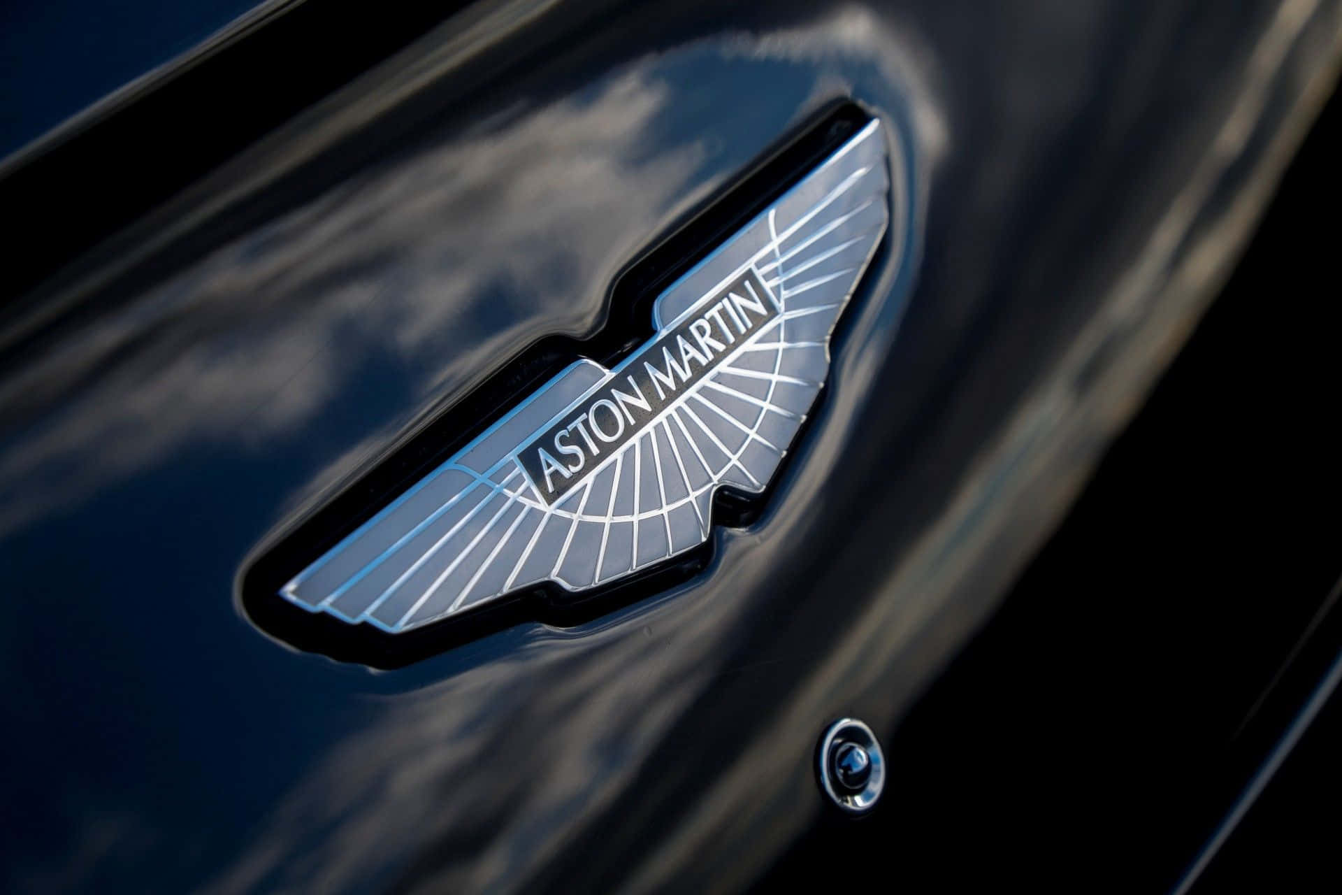 奧斯頓馬丁 桌布 壁紙 — 特寫：閃亮銀色Aston Martin翼形標誌，位於反射陰天雲層的光澤黑色引擎蓋上。