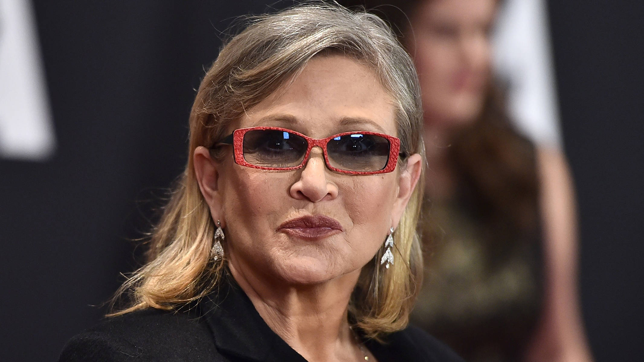 Celebrity 桌布 壁紙 — 特寫一位灰髮女性，戴著大膽的紅色圖案太陽眼鏡和垂墜耳環，背景是深色模糊的。