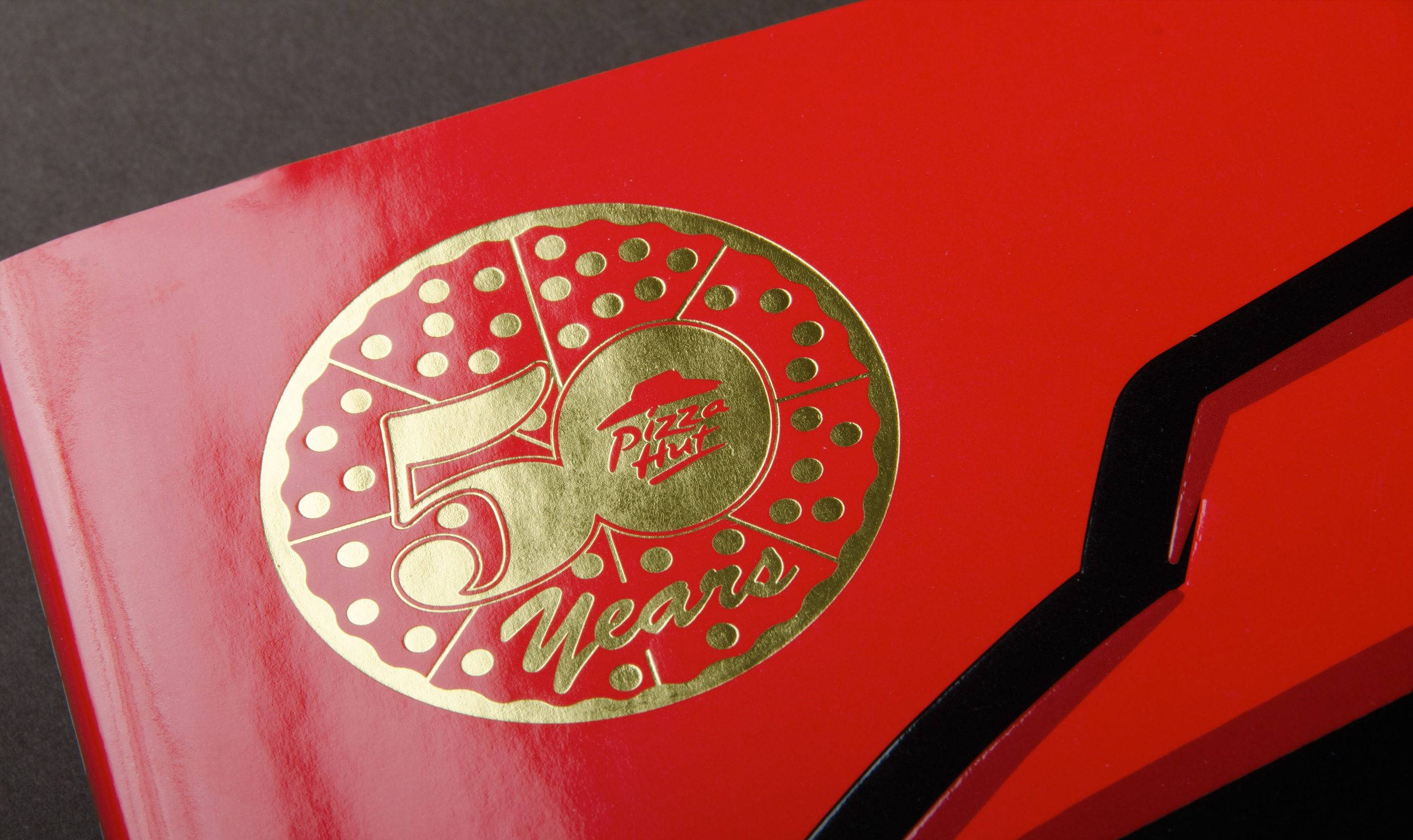 Hut 桌布 壁紙 — 特寫一個亮面紅色盒子，上面有金色箔紙Pizza Hut 50 Years標誌，附有披薩片設計。