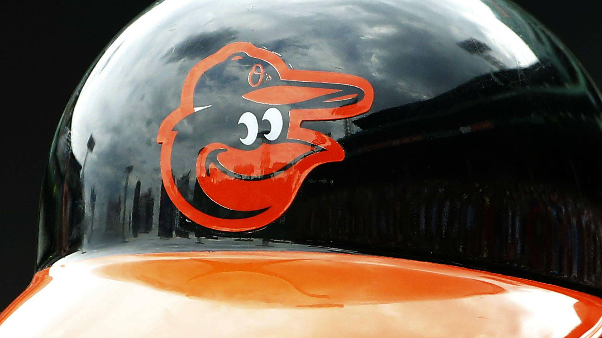 Orioles 桌布 壁紙 — 特寫一個亮黑色棒球頭盔，上面有明亮的橙色卡通黃鸝標誌，反射著多雲的天空。