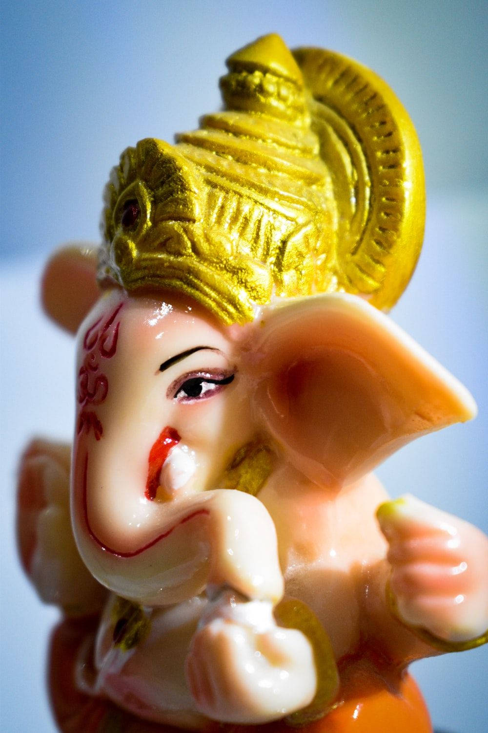 ganpati HD 桌布 壁紙 — 特寫一個帶有金色皇冠和紅色標記的光澤粉紅色象神雕像，背景是柔和的藍色。