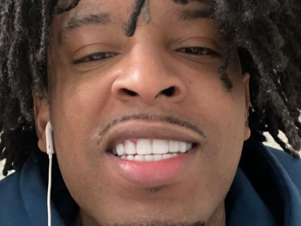 21 Savage 桌布 壁紙 — 特寫一個微笑的男人，深色辮子，白色耳塞，穿著藍色連帽衫。