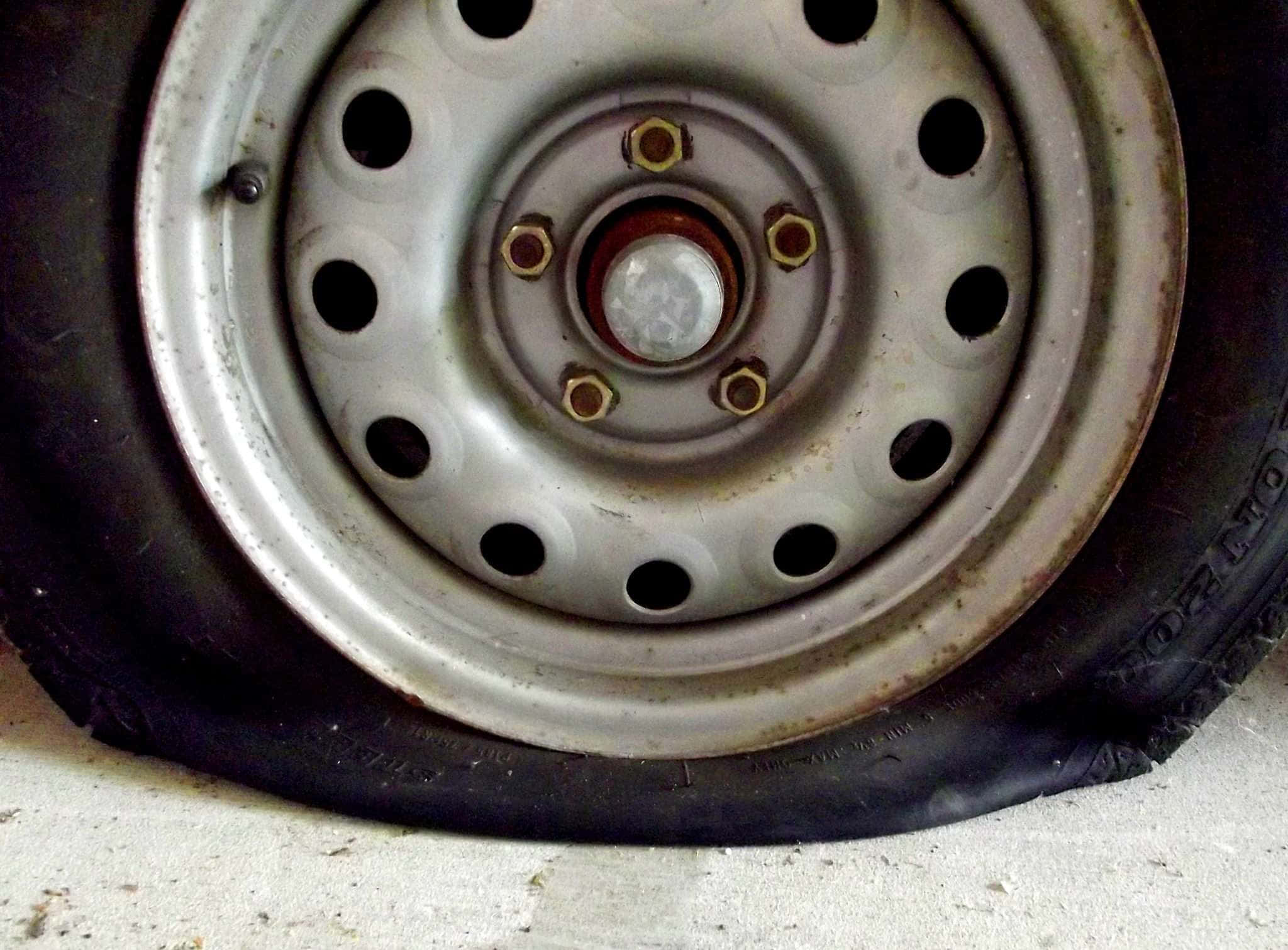 Wheel 桌布 壁紙 — 特寫一個磨損的銀色輪圈，帶有生鏽的螺栓和一個扁平的黑色輪胎，擱在混凝土地板上。