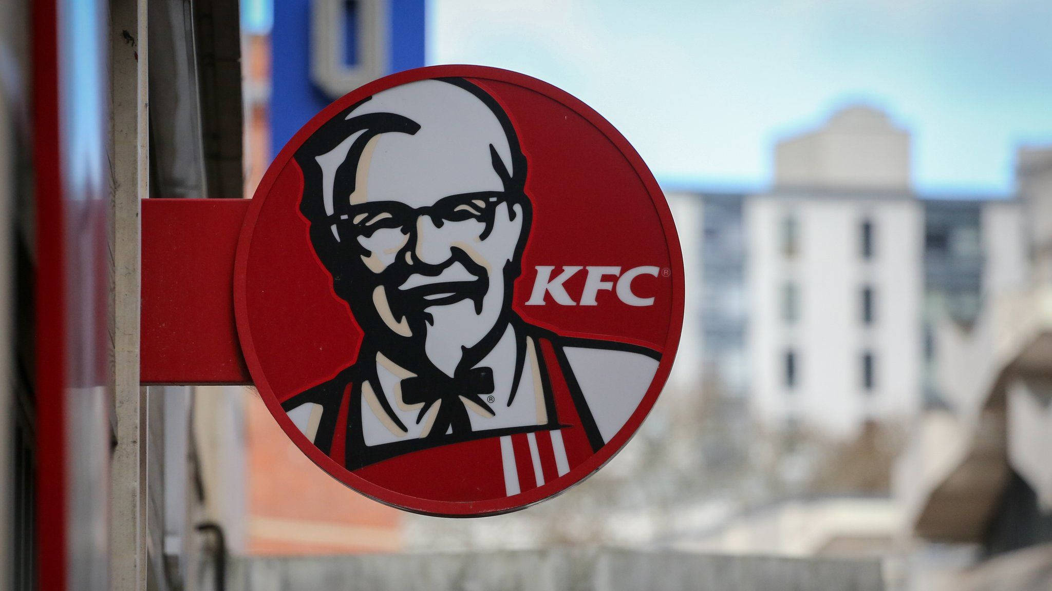 Kfc 桌布 壁紙 — 特寫一個紅色圓形KFC招牌，上面有桑德斯上校的笑臉，背景是模糊的城市景觀。